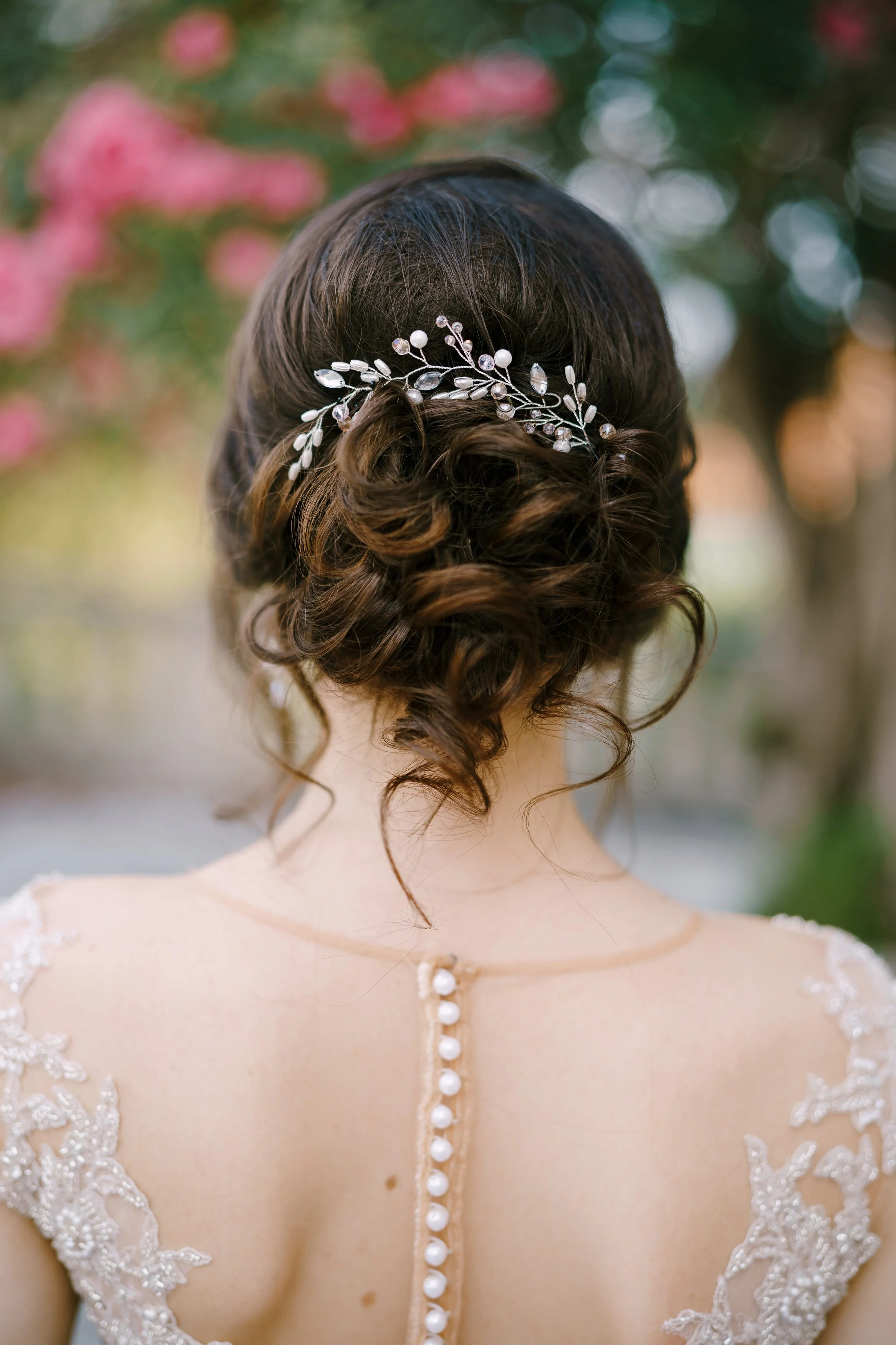 Back view of a woman with dark brown hair styled in loose curls, wearing a decorative hairpiece with pearls and crystals, and a wedding dress with lace detailing and pearl buttons on the back.