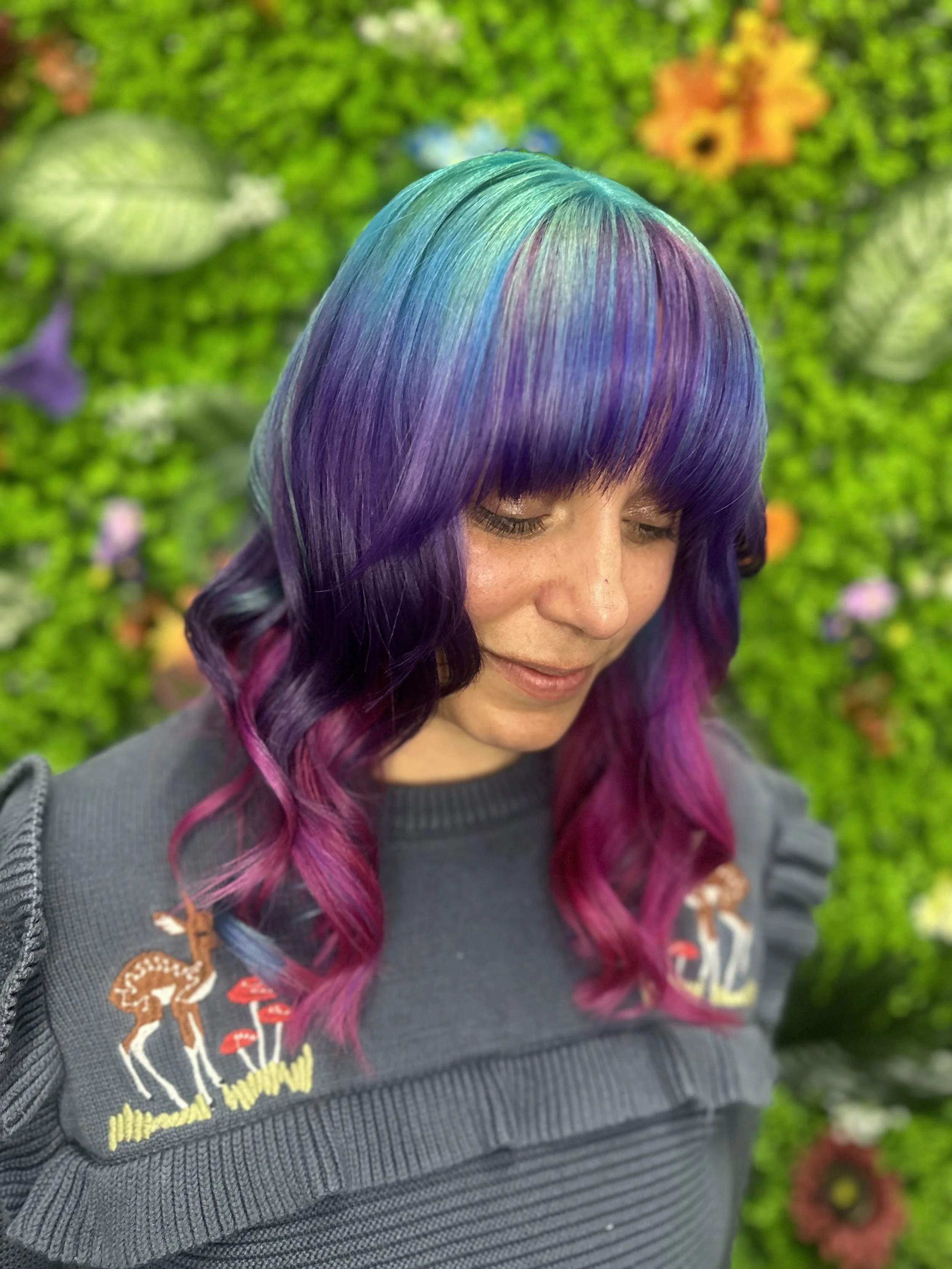Close-up of a woman with long, wavy hair dyed in rainbow colors, with a mix of purple, pink, blue, green, and yellow, against a blurred green floral background.