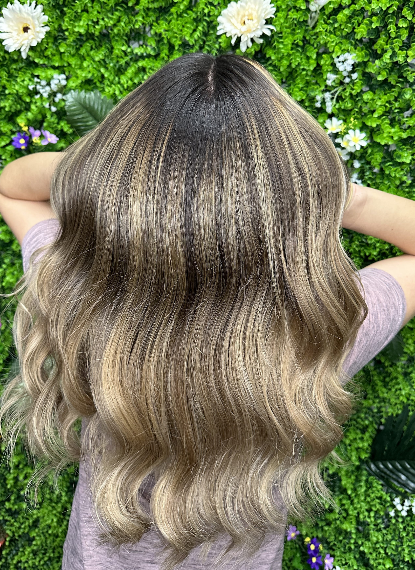 Top view of a person's head with long, wavy, blonde hair against a green leafy background with white, purple, and yellow flowers.