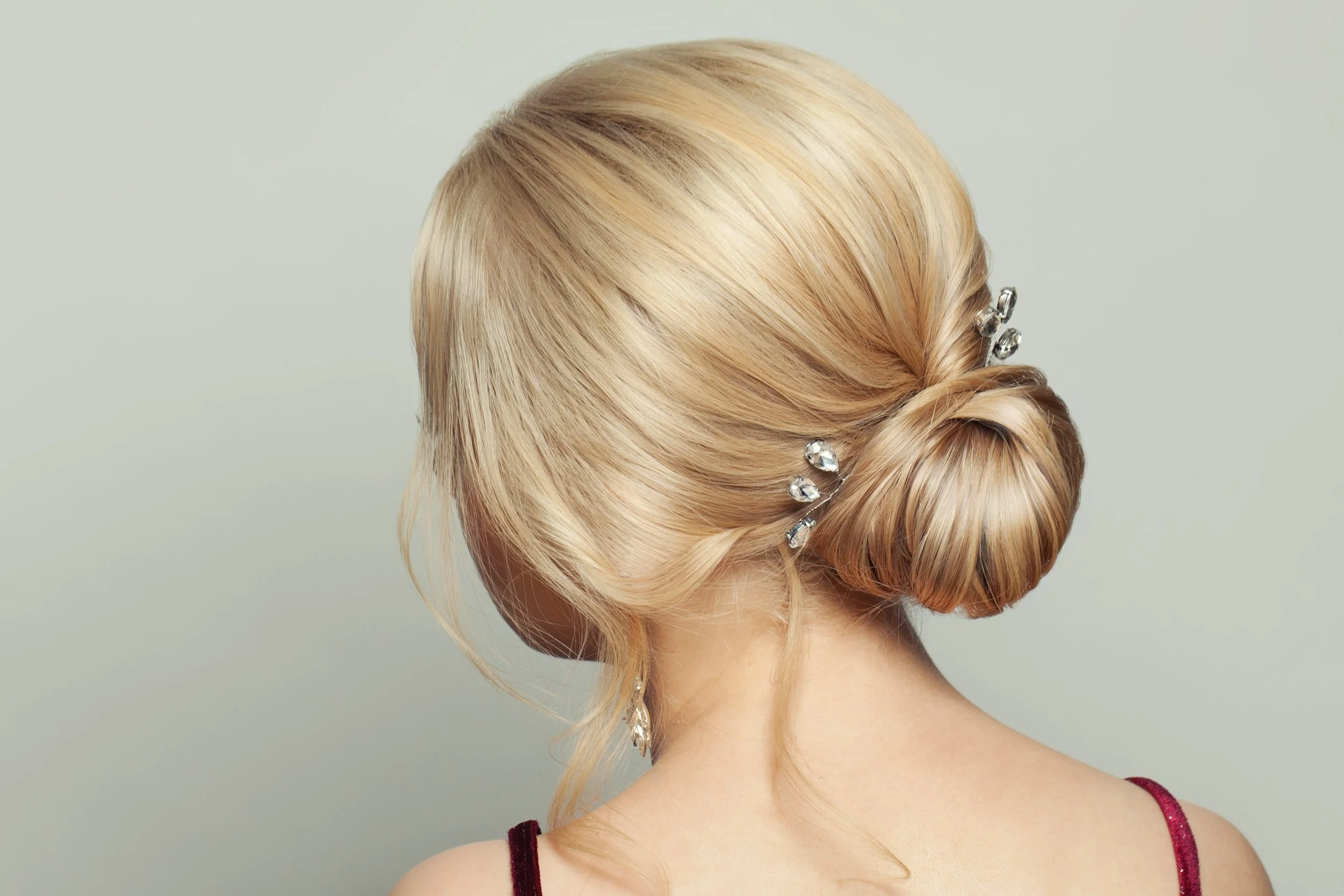 A woman with blonde hair styled in an elegant low bun, decorated with crystal hairpins, and wearing a dark red dress with thin straps, against a light background.
