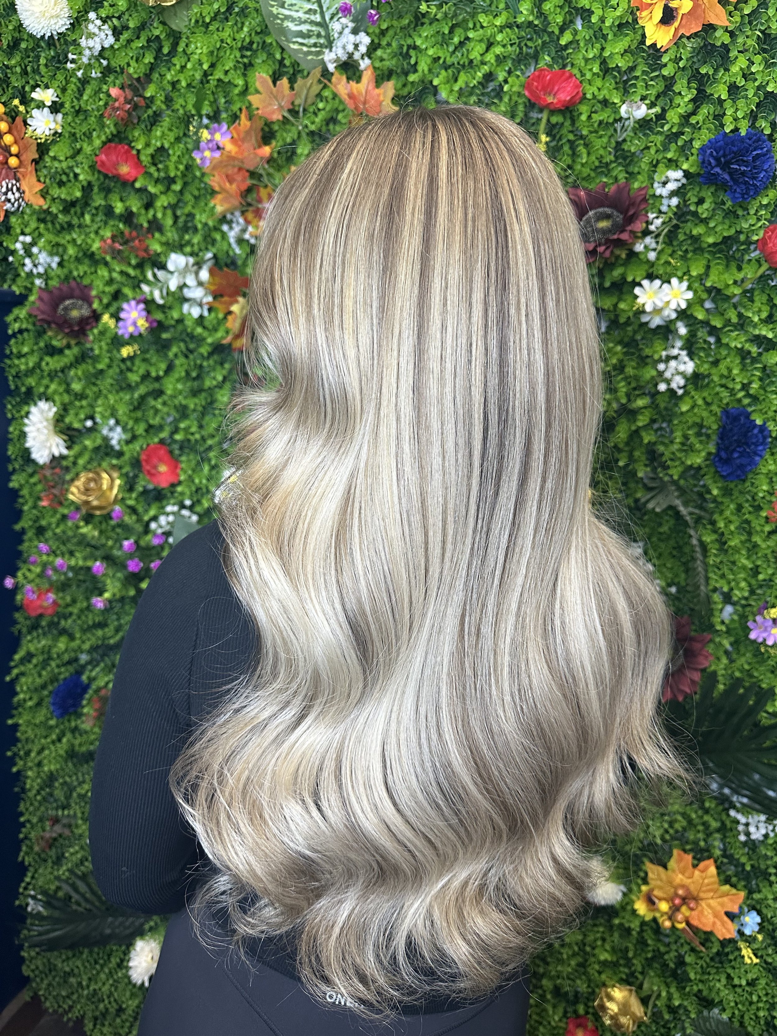 Back of a woman's head with wavy blond hair, standing in front of a green floral and plant wall with colorful flowers and leaves.