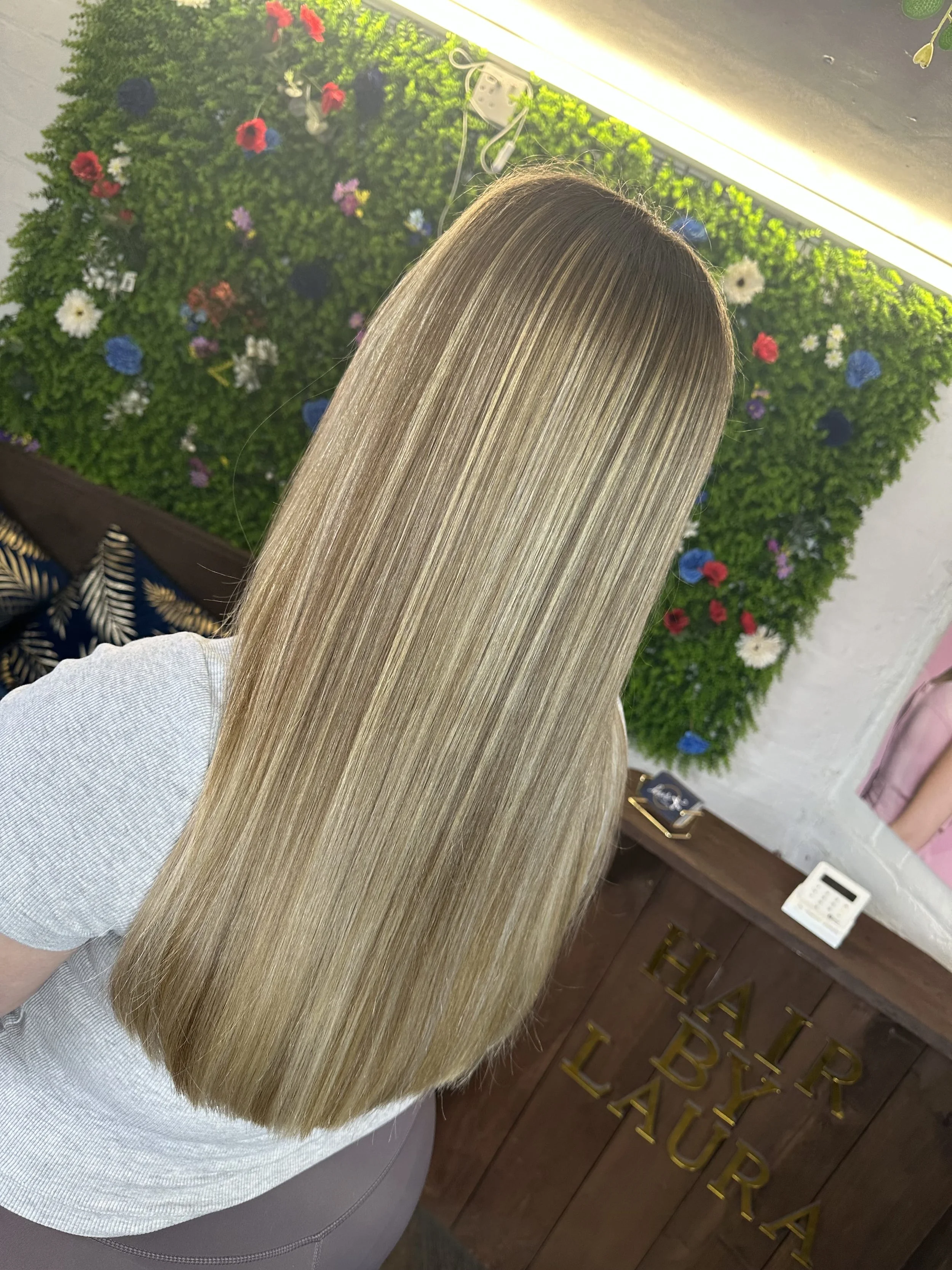 Blonde woman with straight hair standing with her back to the camera in a salon, in front of a green wall decorated with colorful flowers and a sign that reads 'HAIR UP'