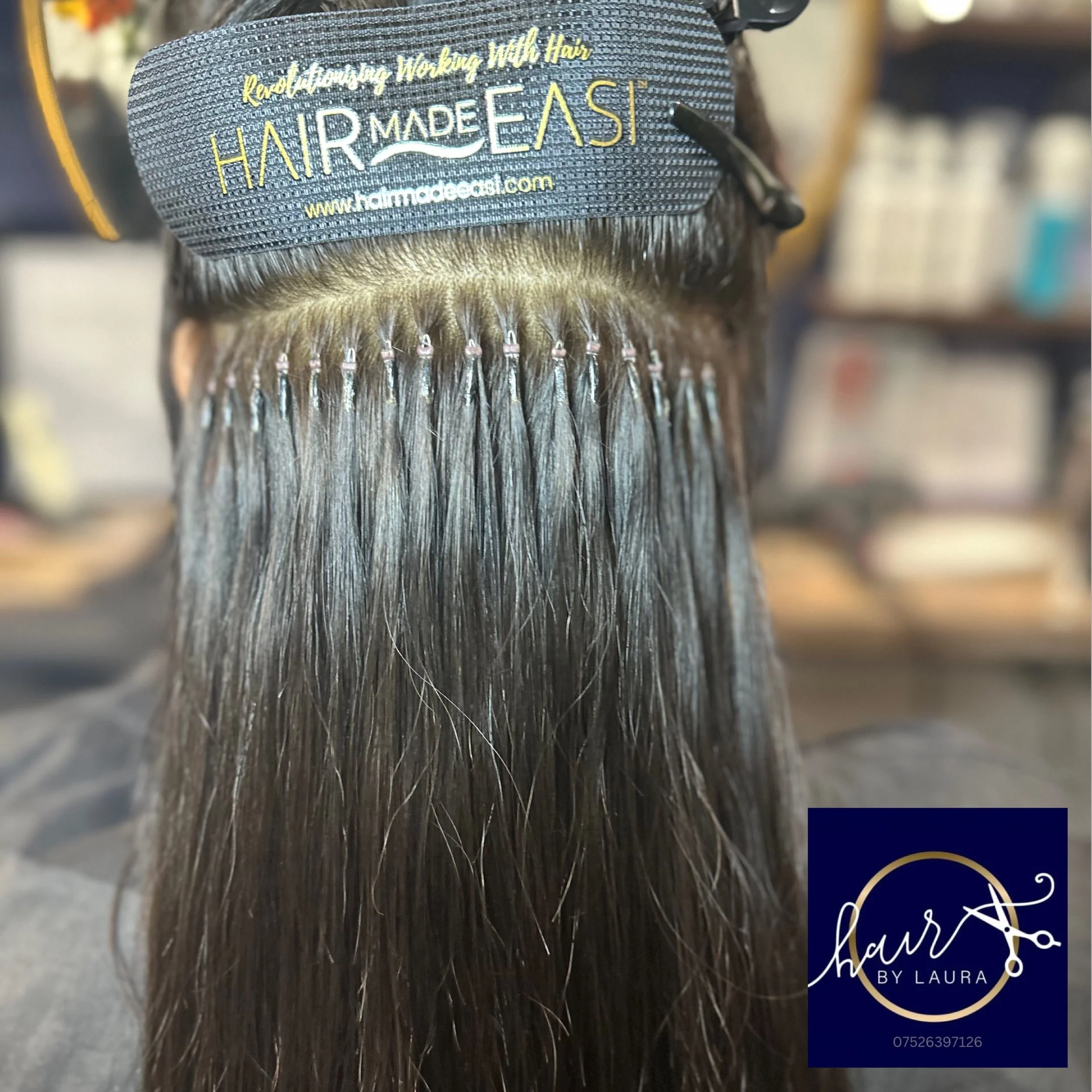 Close-up of a hair extension clip-in with a fabric tag that reads 'Kendall, Working with Hair, Hair Made Easy, www.hairmadeeasy.com' on a person's head with straight dark hair.