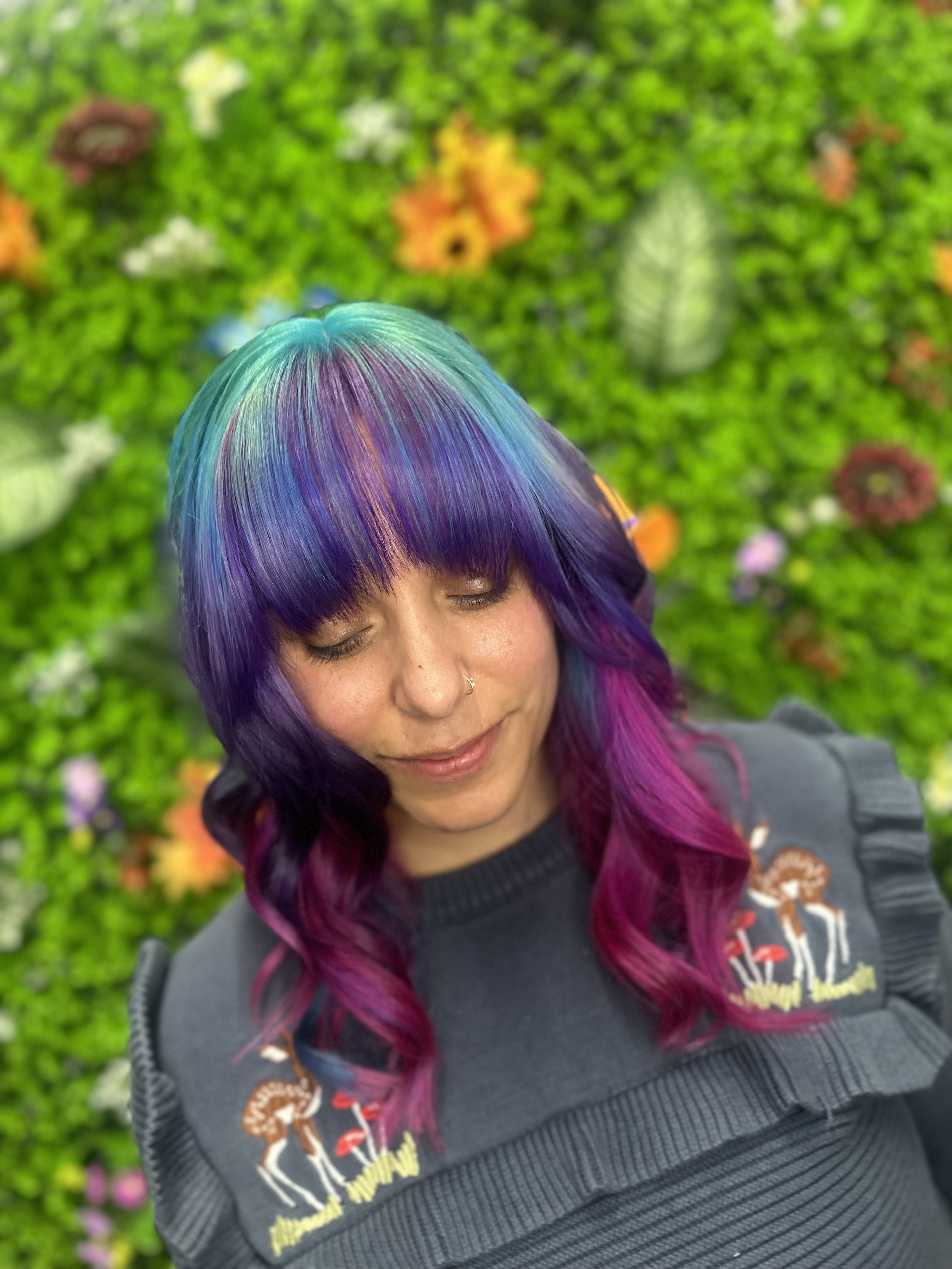 A woman with multicolored hair in shades of turquoise, purple, and pink, with closed eyes, wearing a dark jacket with embroidered patches, standing in front of a green floral wall.