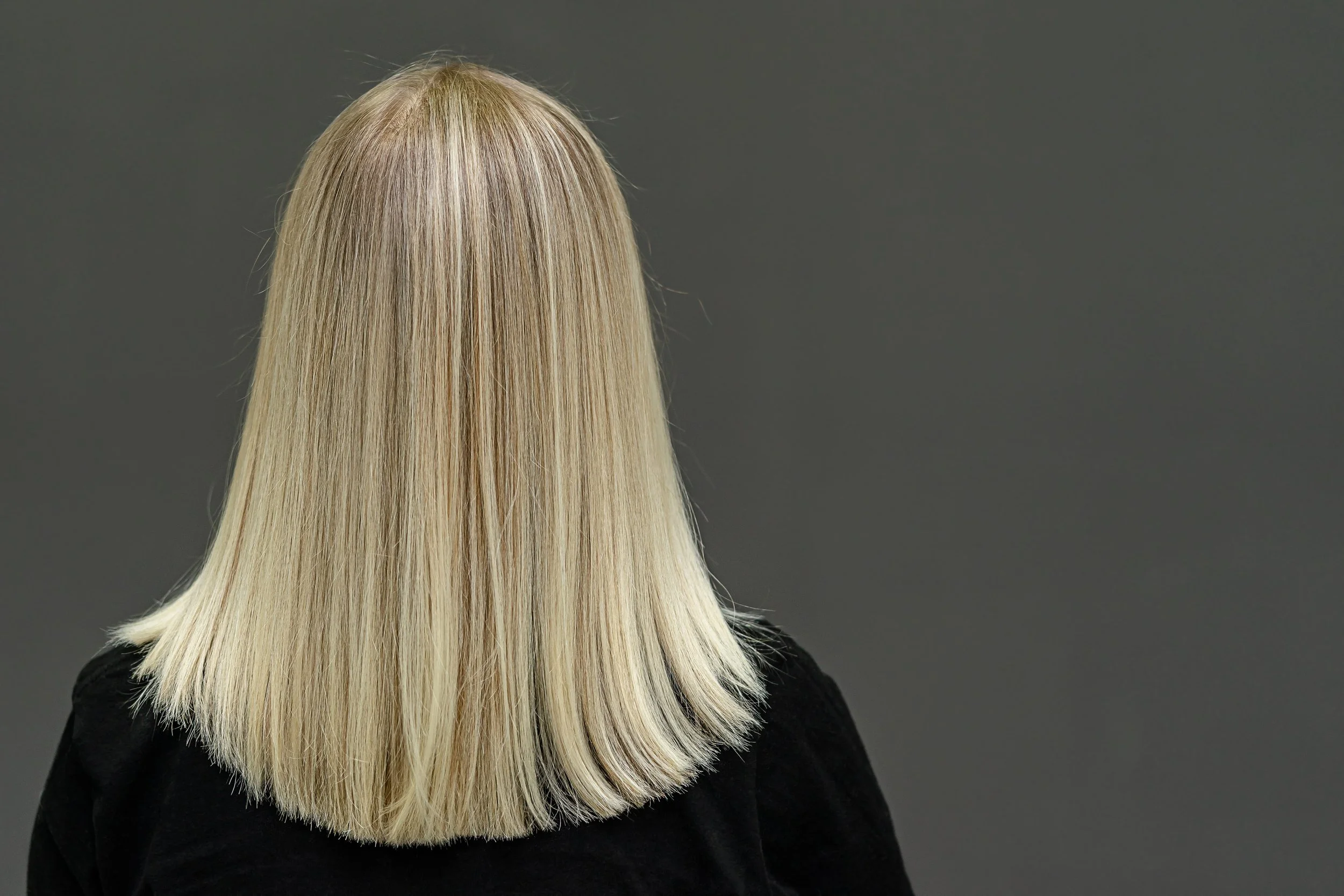 Back view of a woman with straight, platinum blonde hair wearing a black top, standing against a plain gray background.