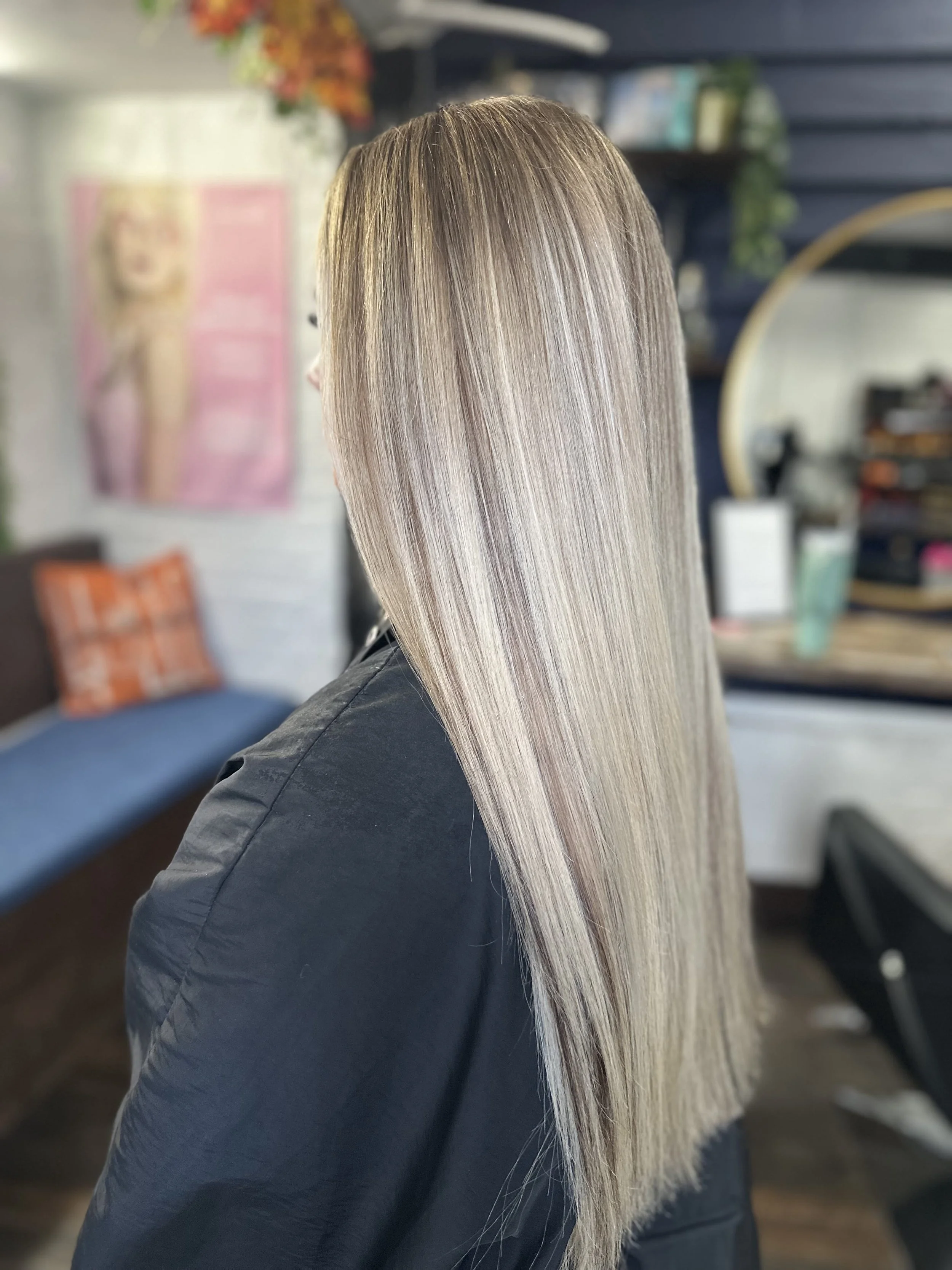 Person with long, straight, blonde hair sitting in a salon with shelves and decorations in the background.