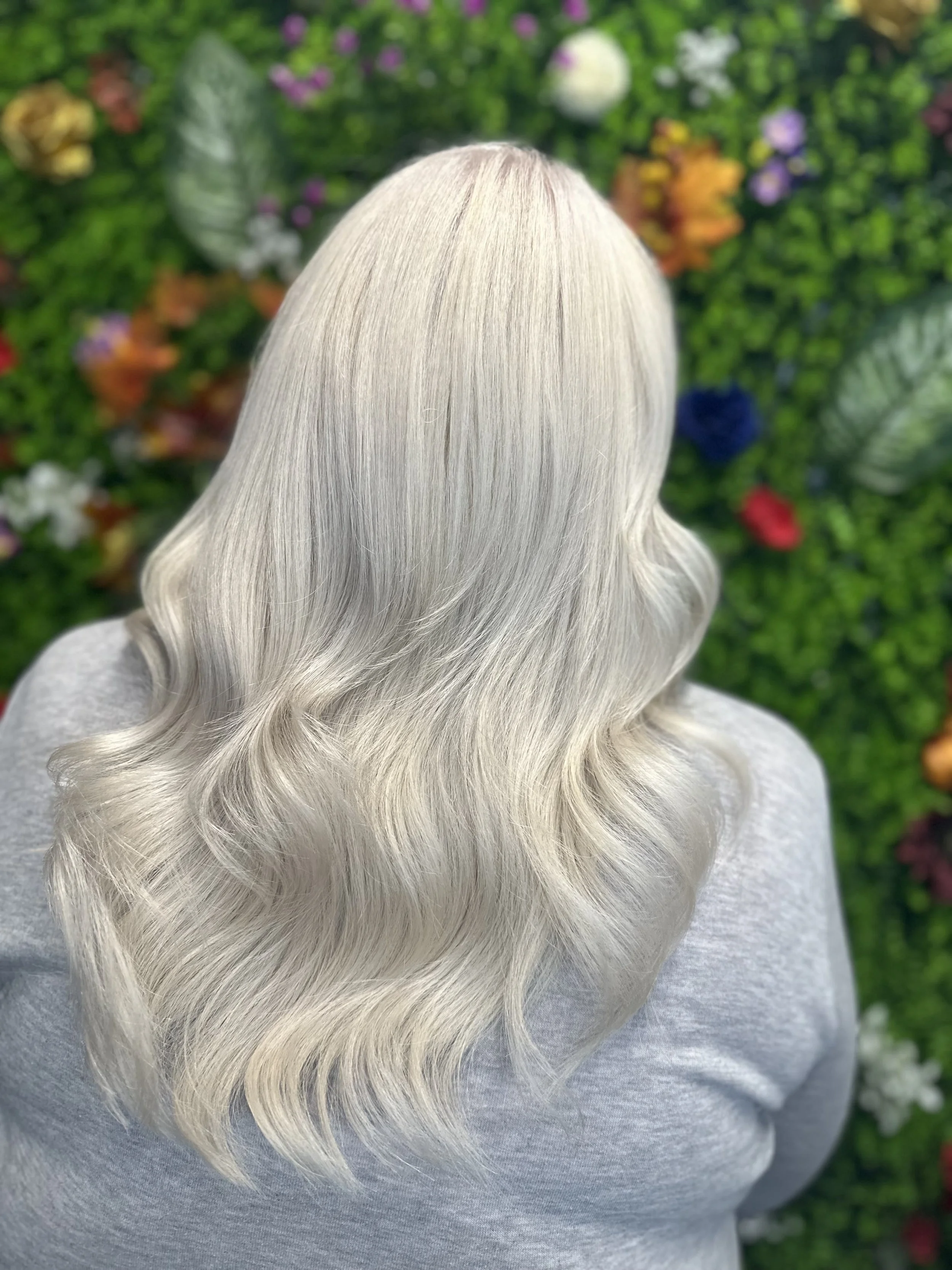 Close-up of a person with long, wavy, platinum blonde hair, standing outdoors against a garden wall with various green plants and colorful flowers.