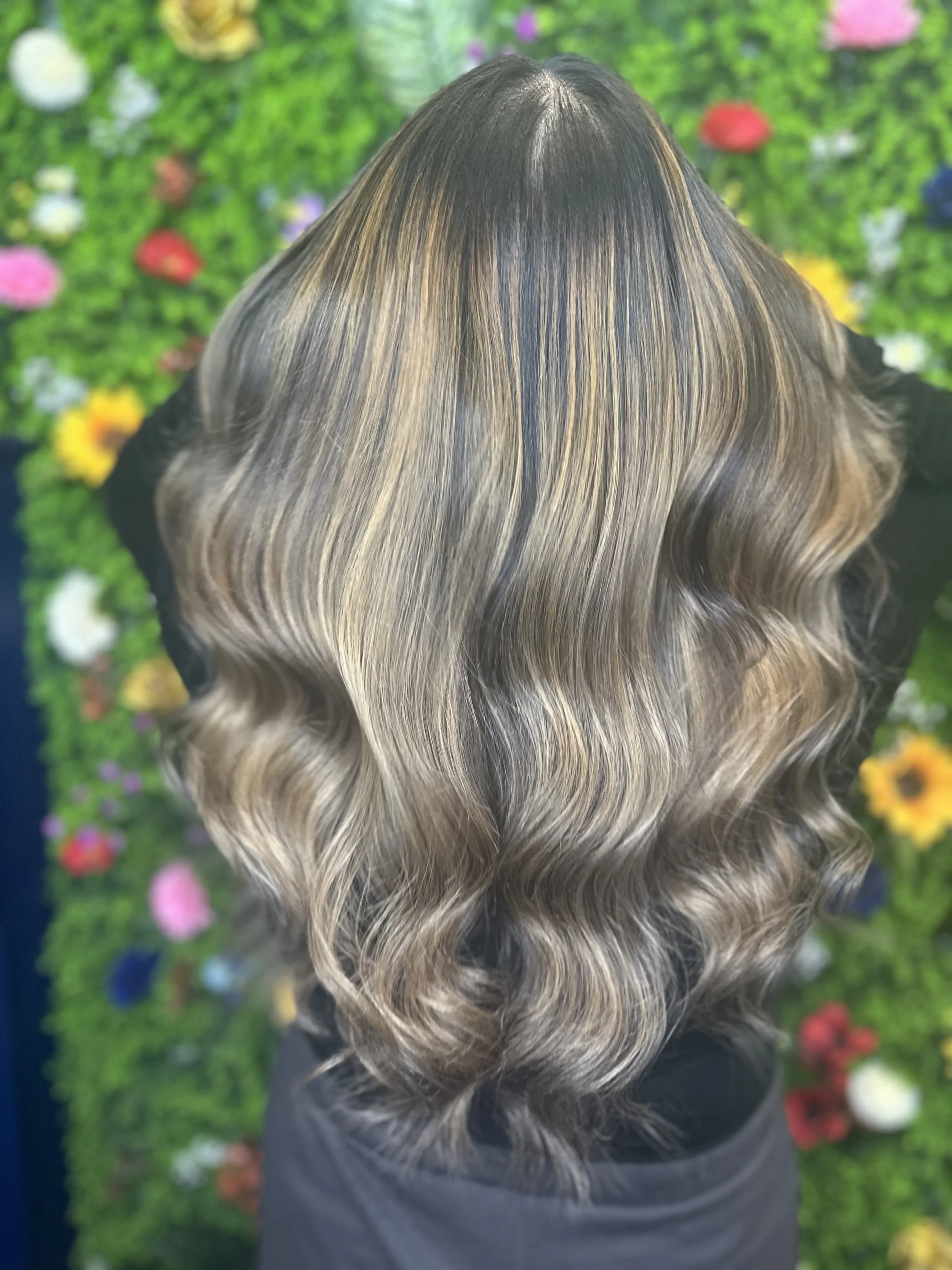 Back view of a person with long, wavy hair with blonde highlights, standing in front of a green, colorful, floral background.