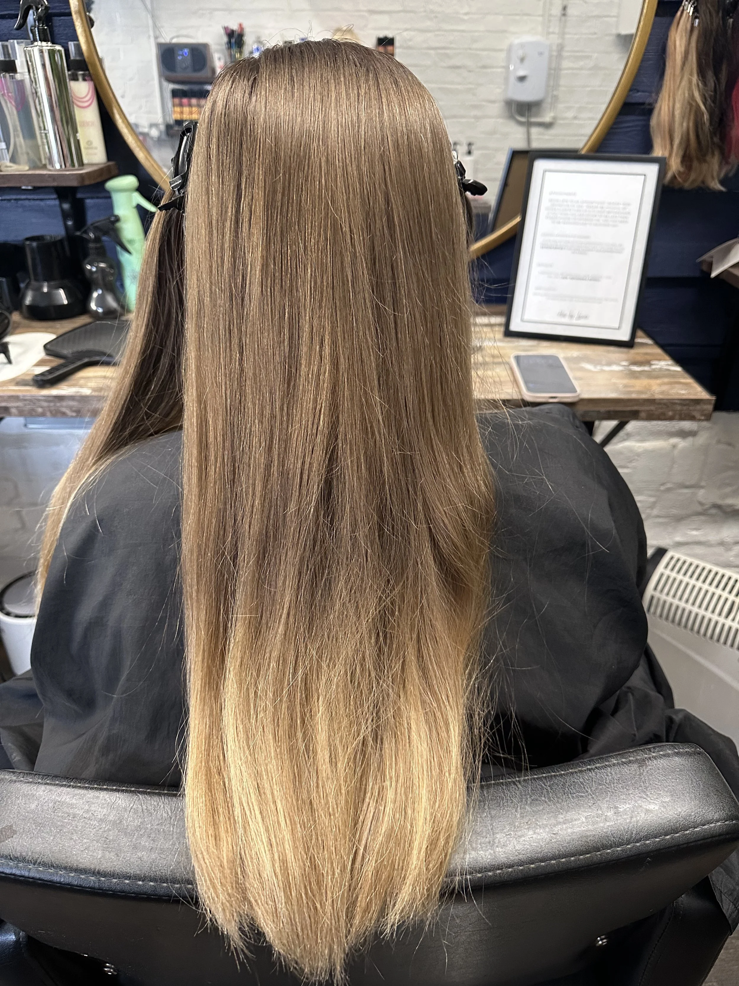 The image shows the back of a person's head with long, straight, light brown or blonde hair in a salon setting. There is a mirror and various hair products in the background.