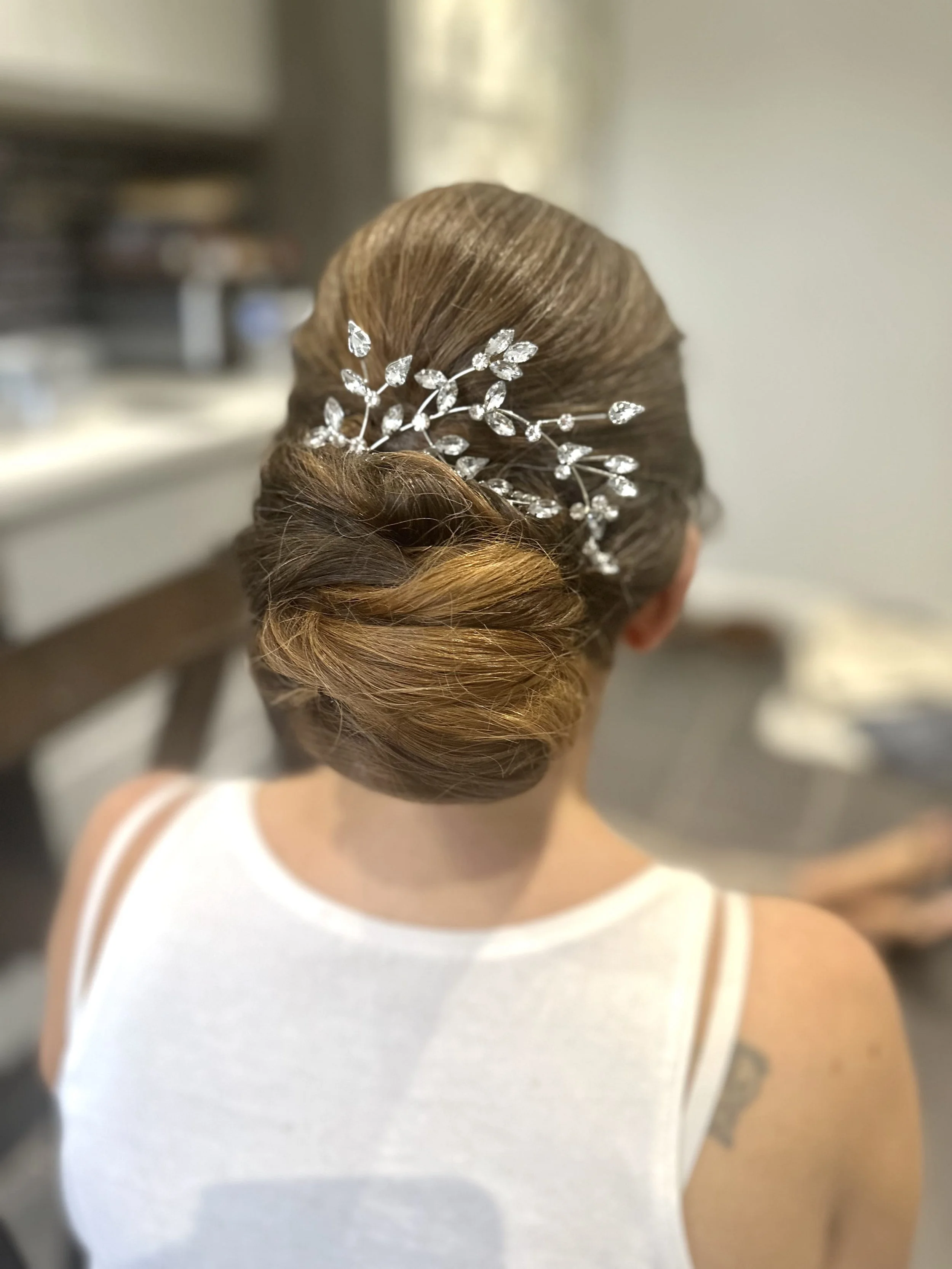 Back of a woman with styled brown hair in an updo, decorated with a silver leaf hair accessory, wearing a white sleeveless dress, standing indoors near a counter.