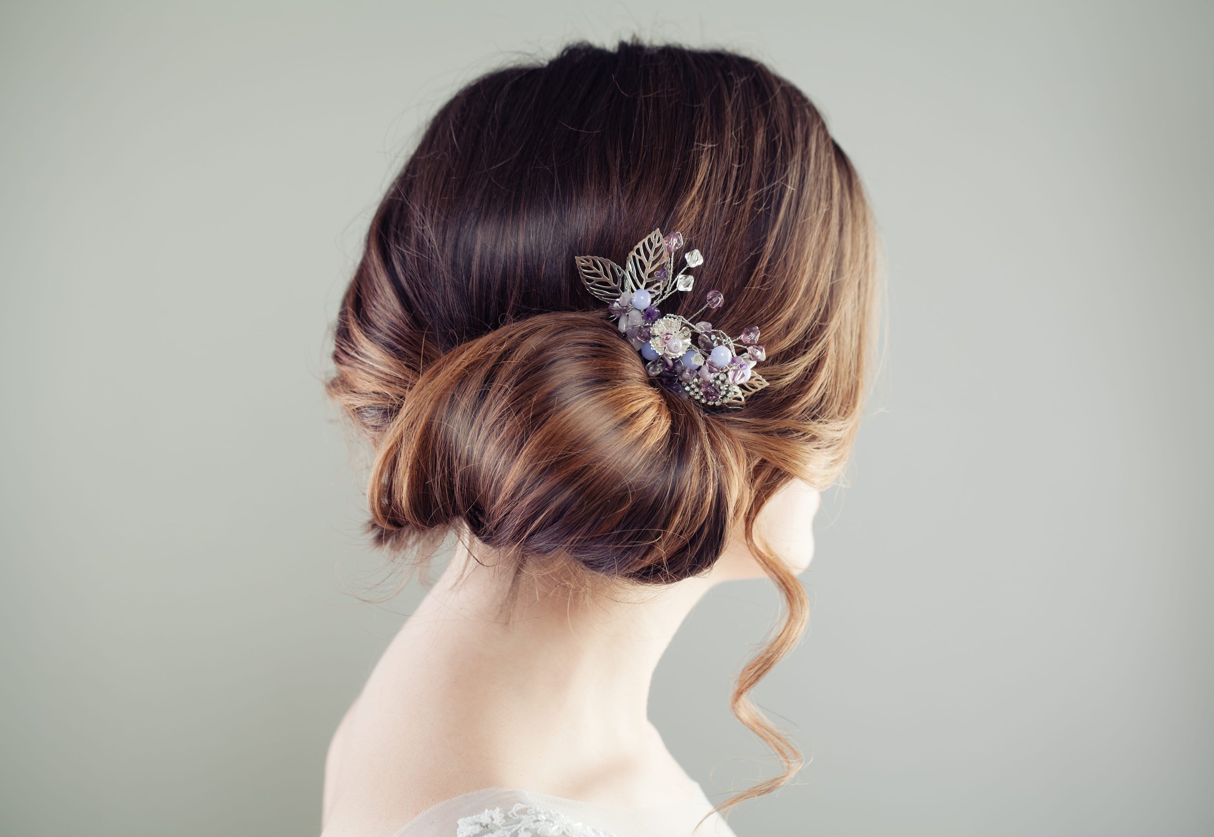 Back view of a woman with elaborately styled hair in a low bun, adorned with a decorative hairpiece featuring leaves and small purple flowers, against a plain light background.