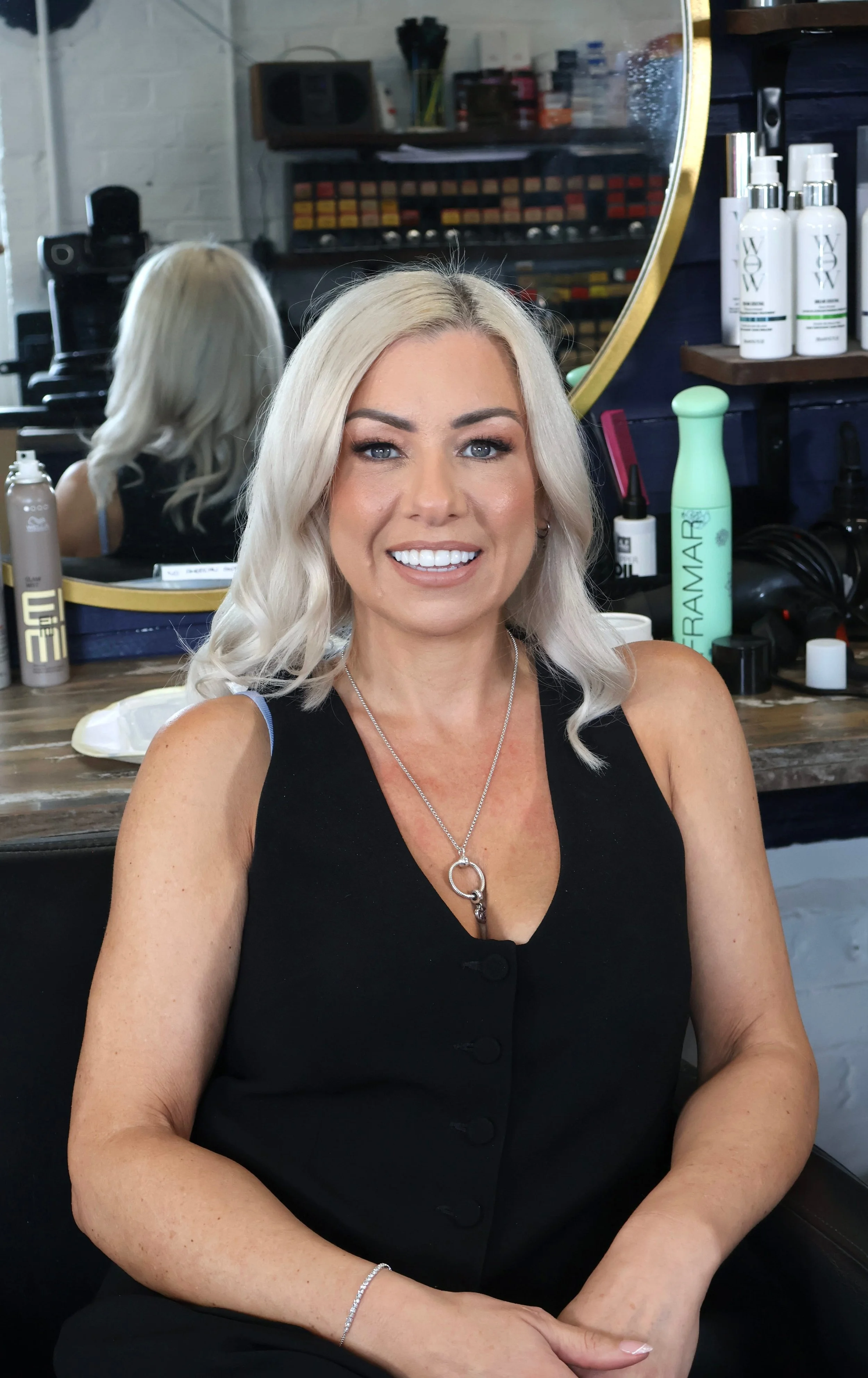 A woman with platinum blonde hair smiling at the camera. She is wearing a black sleeveless top and jewelry, with a salon or studio background including hair products, a mirror, and hair styling tools.