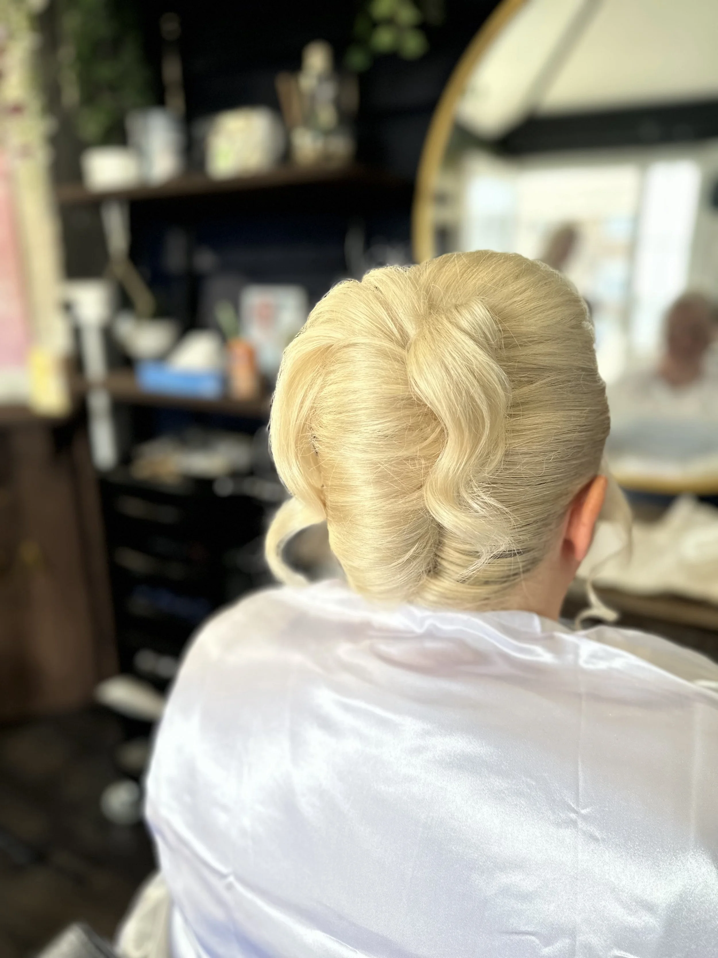 Back of a person with platinum blonde hair styled in an elegant updo, sitting in a salon, wearing a white cape.