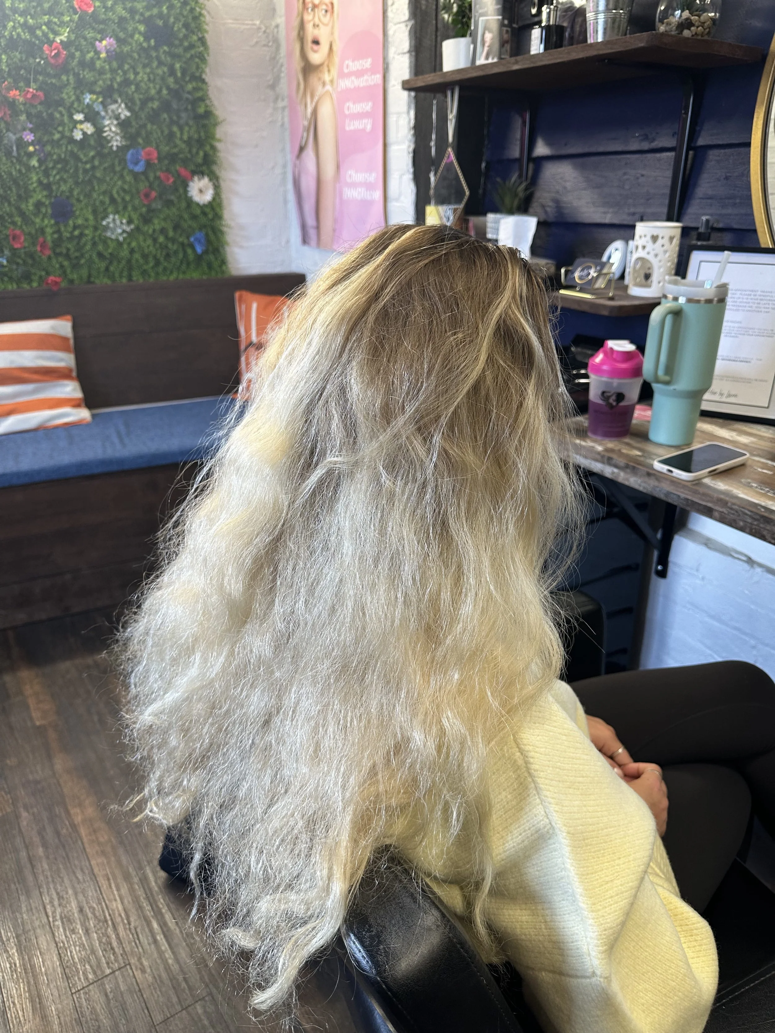 Woman with long, lightly blonde hair sitting in a hair salon, wearing a yellow sweater, with various hair and beauty products on the shelf behind her. The salon has a dark wooden wall and a colorful floral art piece on the wall.