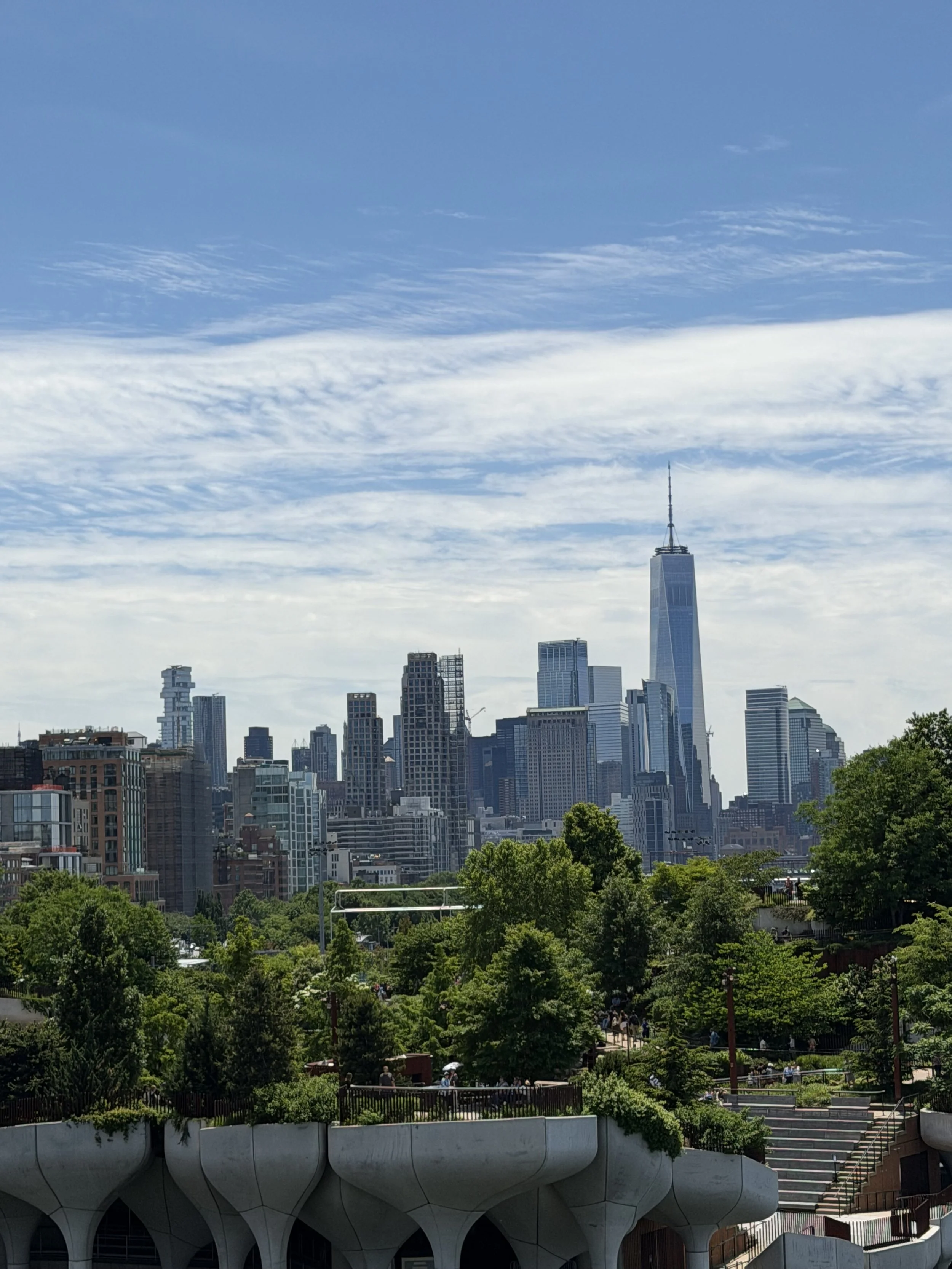 City skyline with tall skyscrapers, including the One World Trade Center, seen from a park with lush trees and a distinctive modern white building with rounded columns in the foreground.
