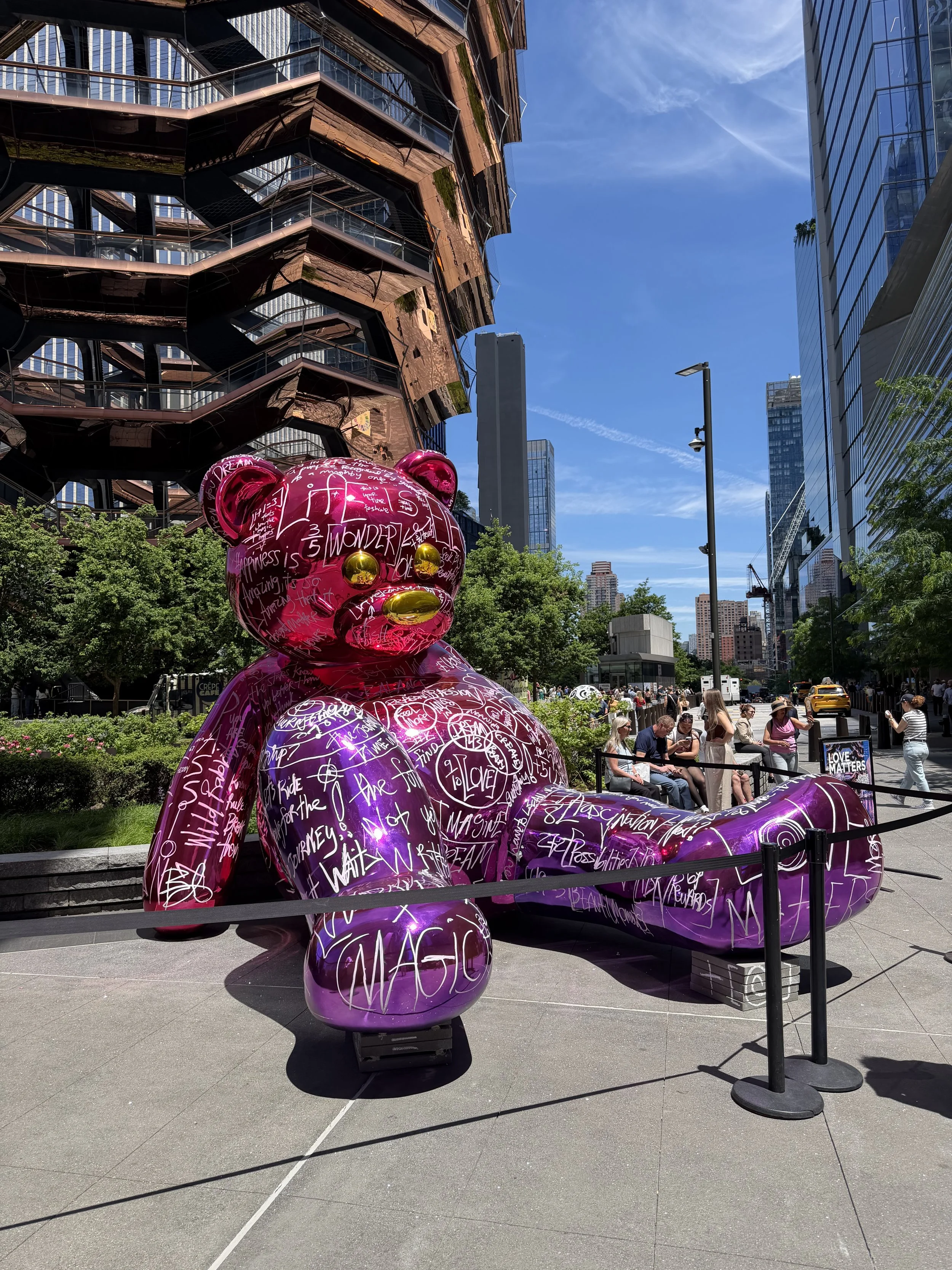 Large pink sculpture of a teddy bear covered in white handwritten words and drawings, situated outdoors with trees, buildings, and a city street in the background.