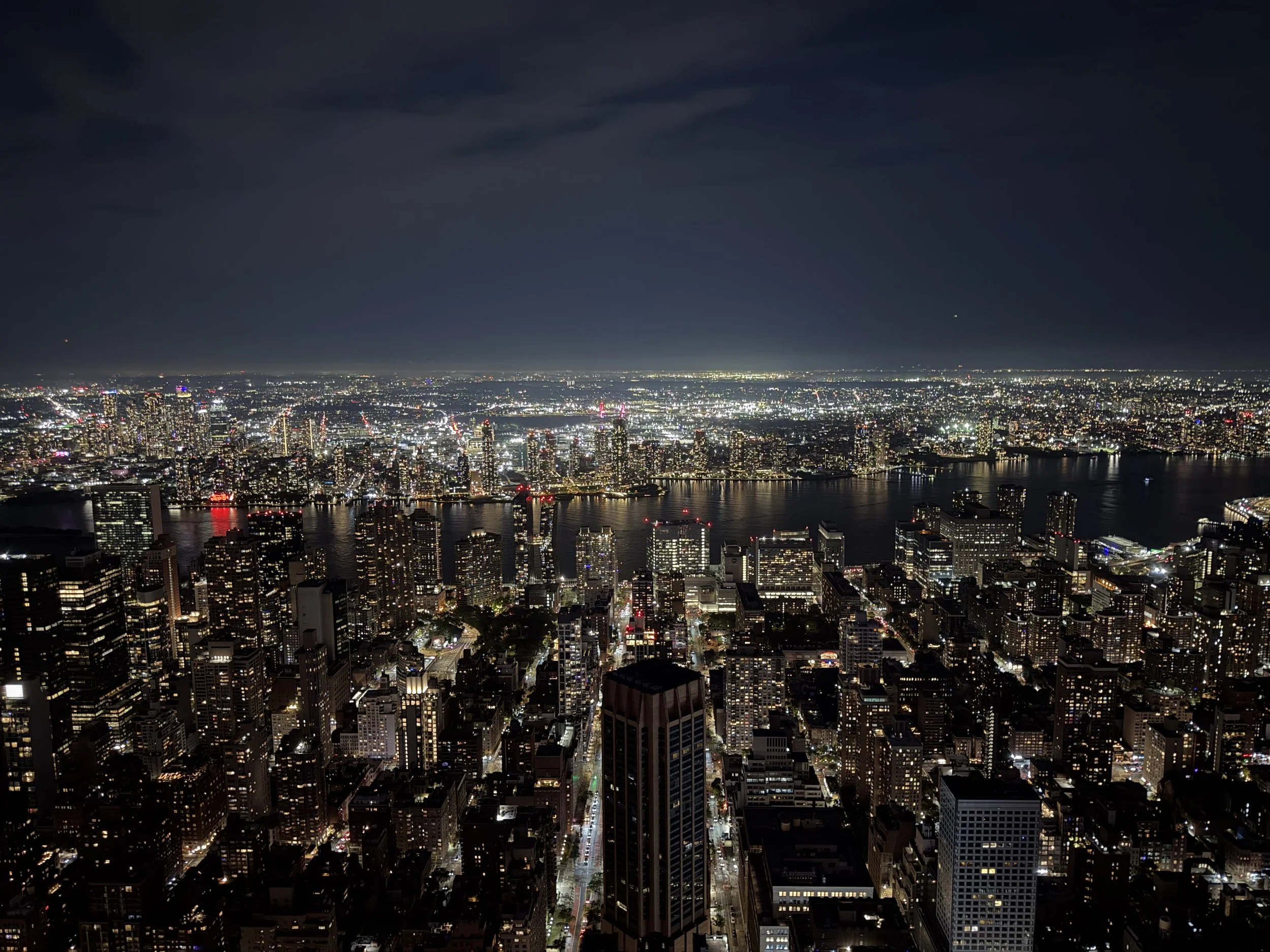 Nighttime aerial view of a city skyline with tall lit-up skyscrapers, a river with illuminated bridges, and a dark sky with scattered clouds.