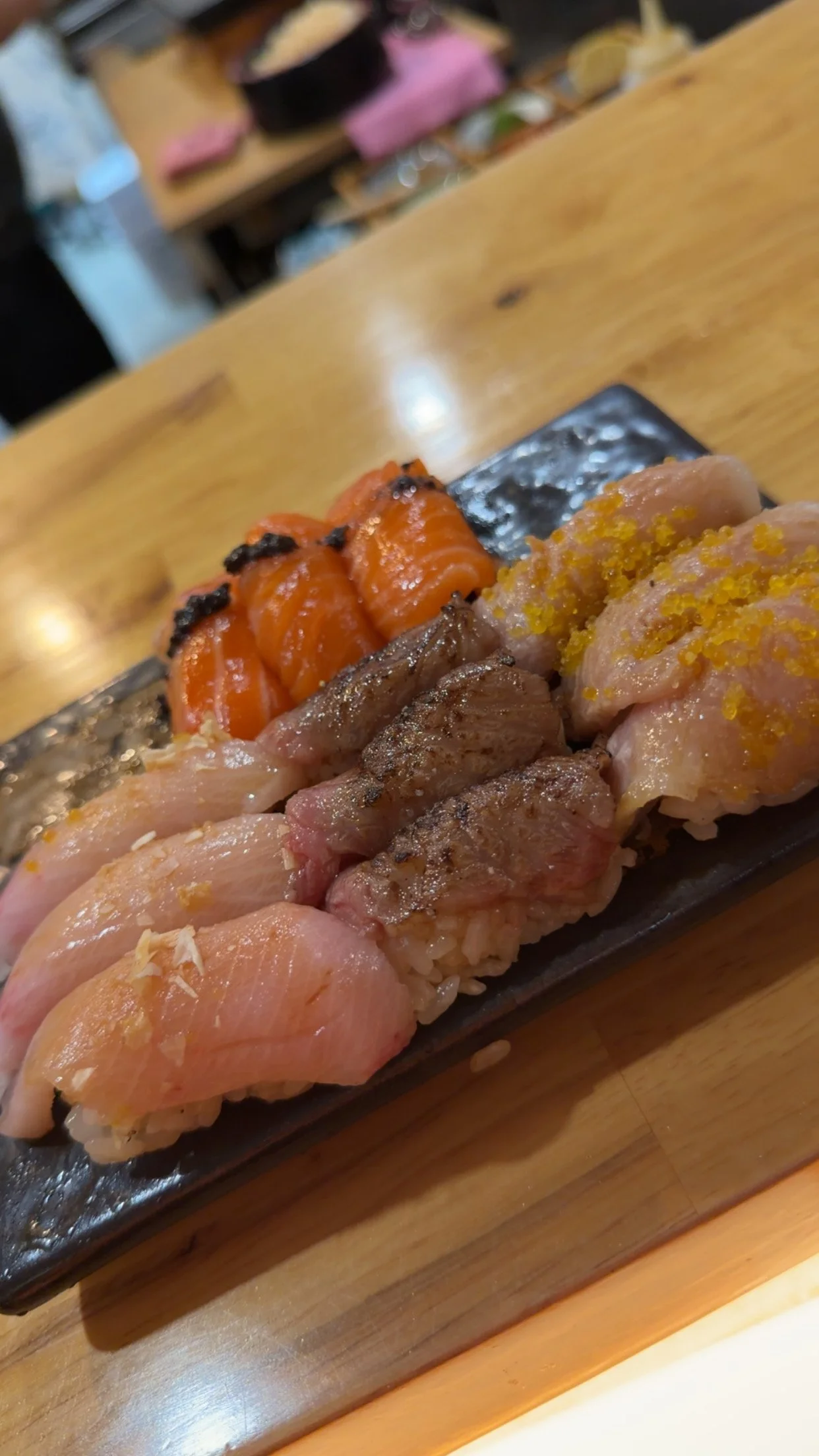 A black rectangular plate with assorted sushi, including salmon, tuna, and other fish, served on a wooden table.