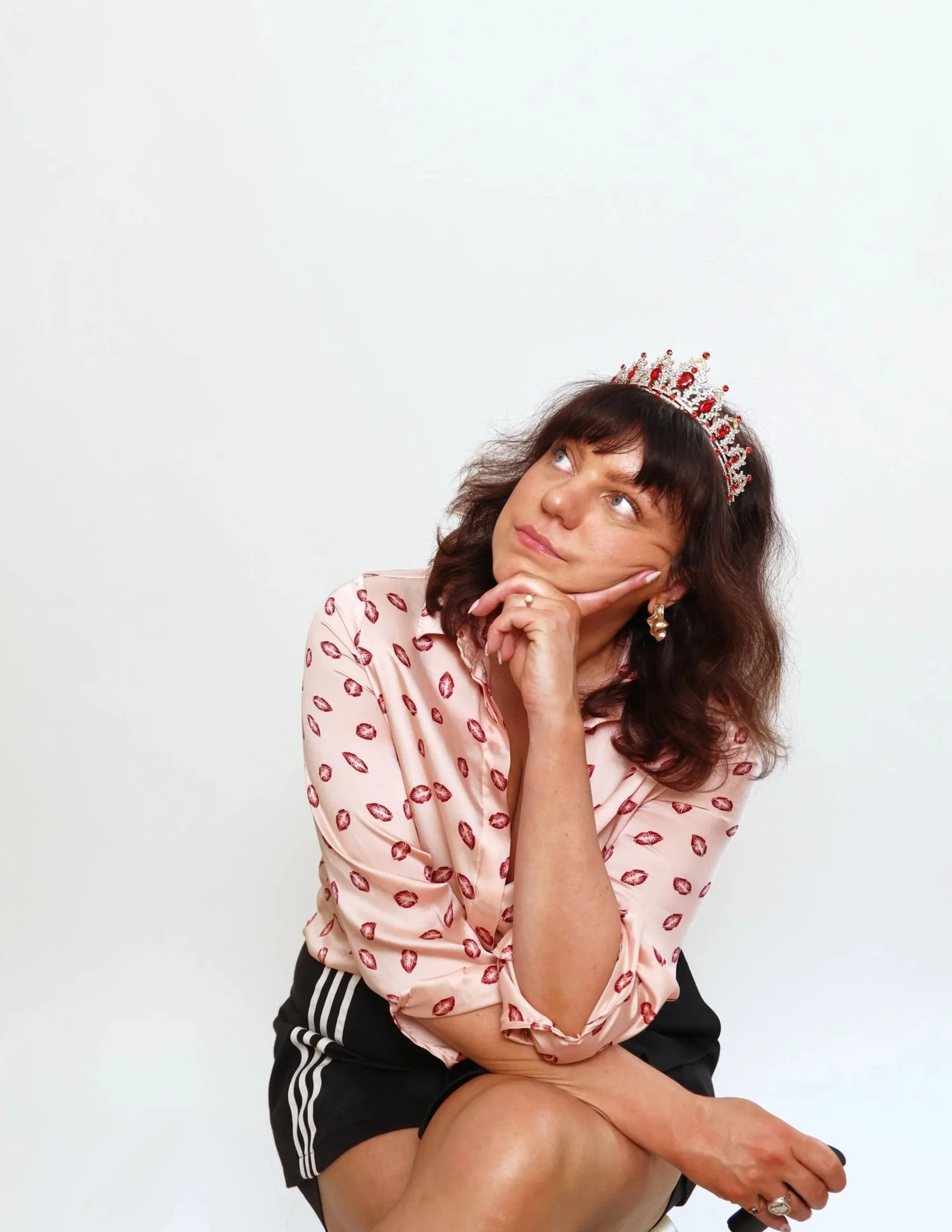 A woman with dark hair wearing a tiara, pink shirt with lips print, and black shorts, sitting with her chin resting on her hand and looking upwards.
