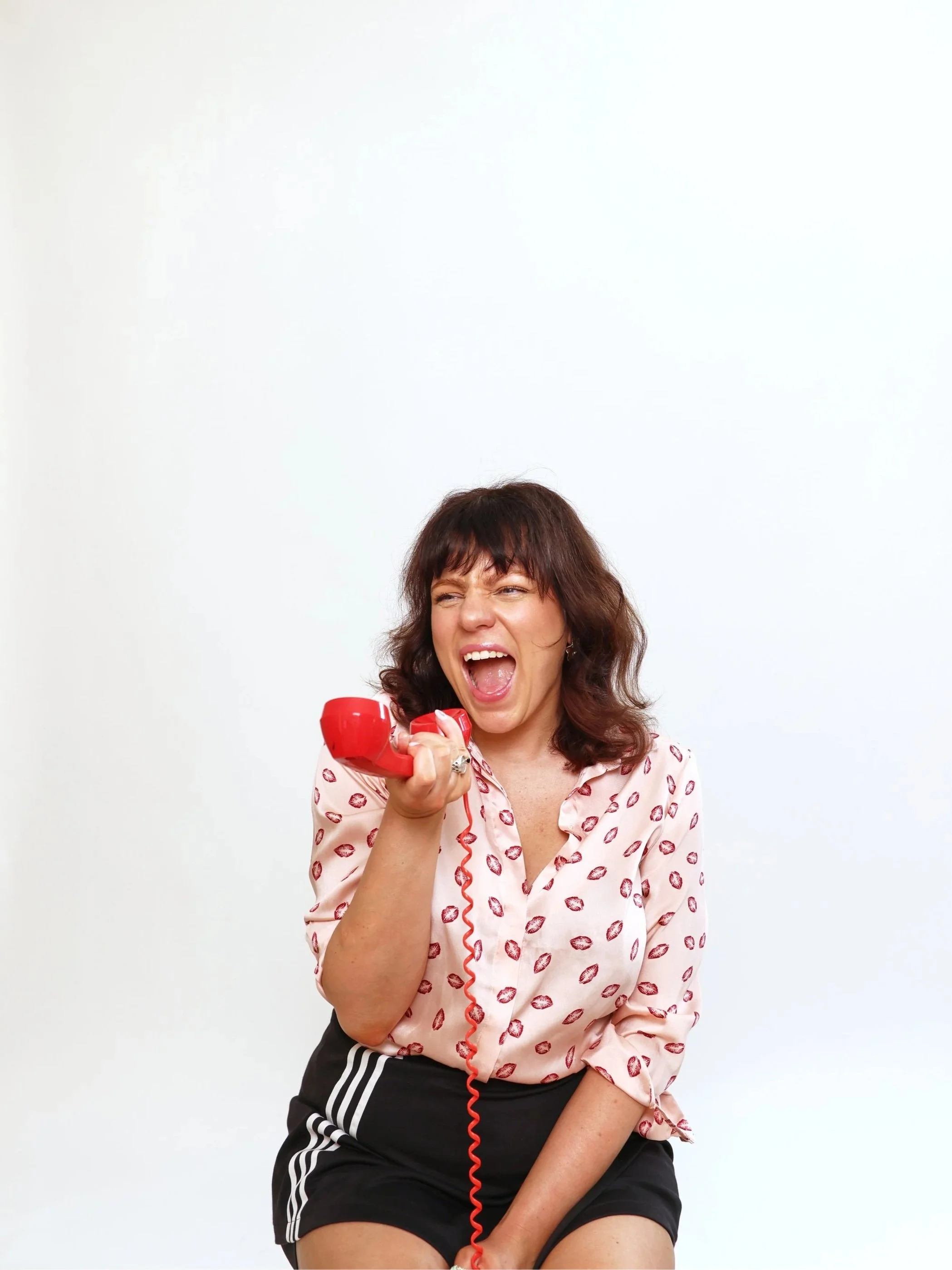 Woman with dark brown hair, wearing a light pink blouse with red lips pattern, holding a red telephone receiver, looking excited