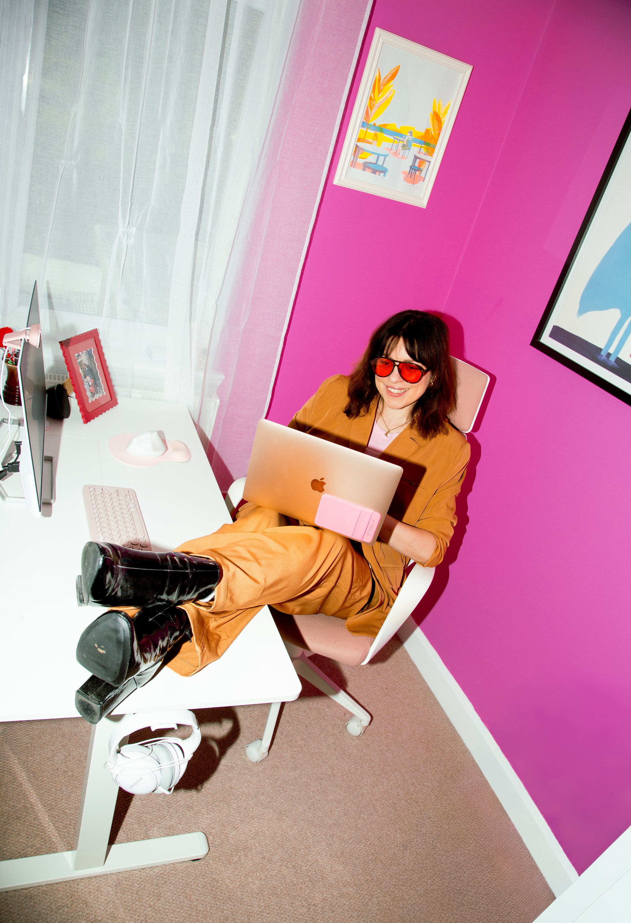 A woman sitting in a pink armchair at a white desk in a colorful room with pink walls, using a silver MacBook that she holds in her lap, wearing red sunglasses and brown clothing, with black boots. The room has white curtains and framed artwork on the walls.