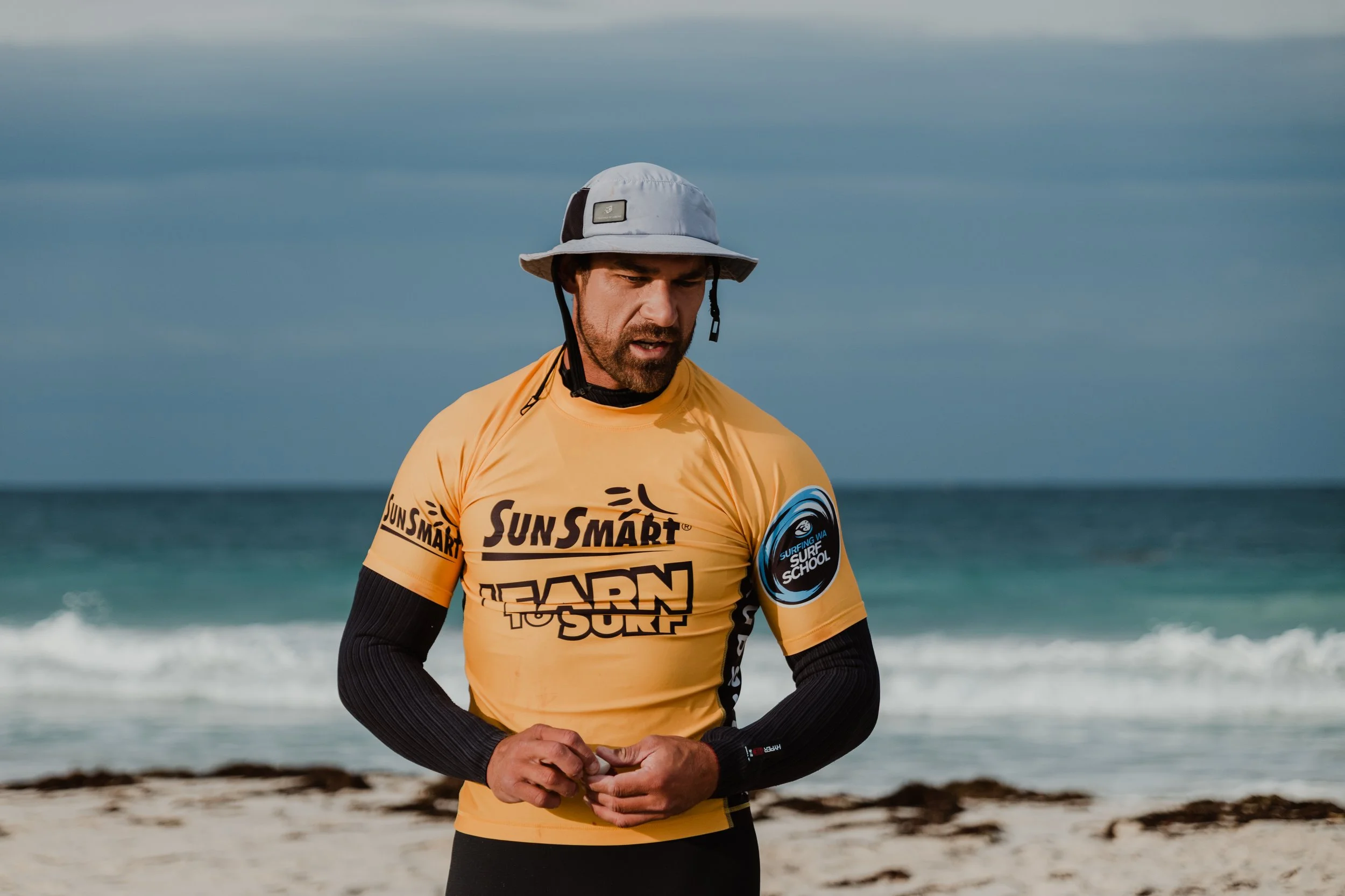 A man wearing a yellow surf school shirt and a wide-brim hat stands on a beach with the ocean in the background.