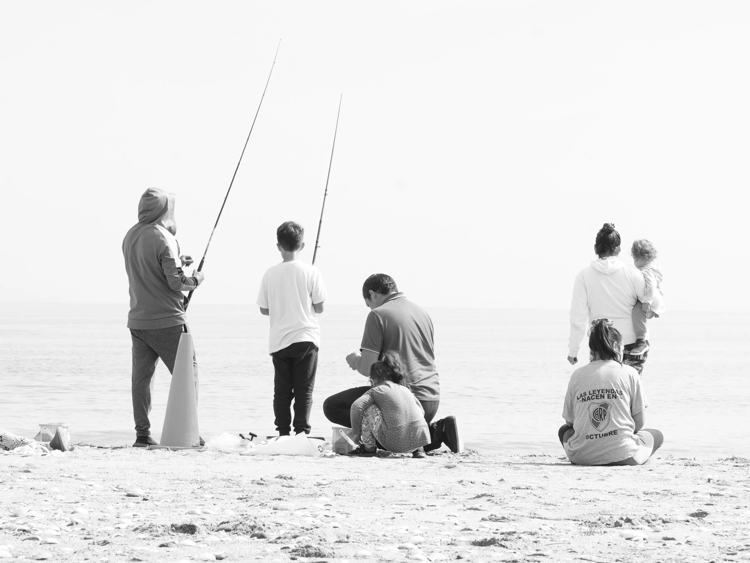 People fishing and relaxing on a beach, some standing, some sitting, with a calm sea in the background.
