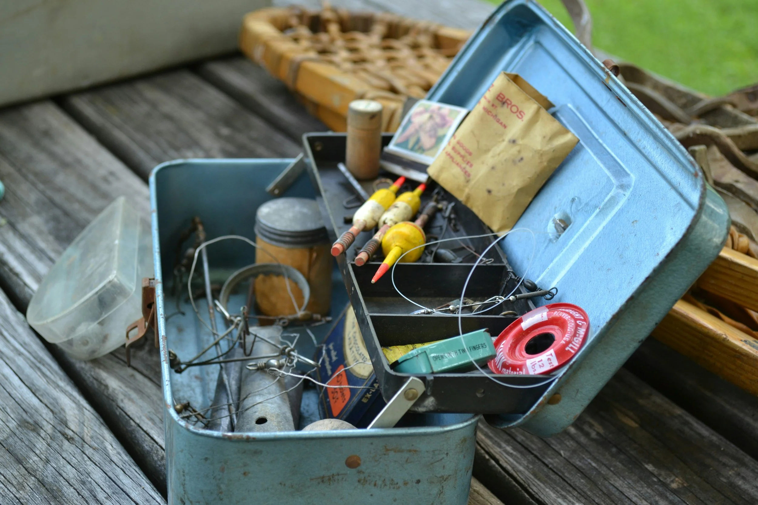 A vintage blue tackle box filled with fishing tackle, including fishing line, small floats, and other fishing accessories, sits on a weathered wooden surface outdoors.