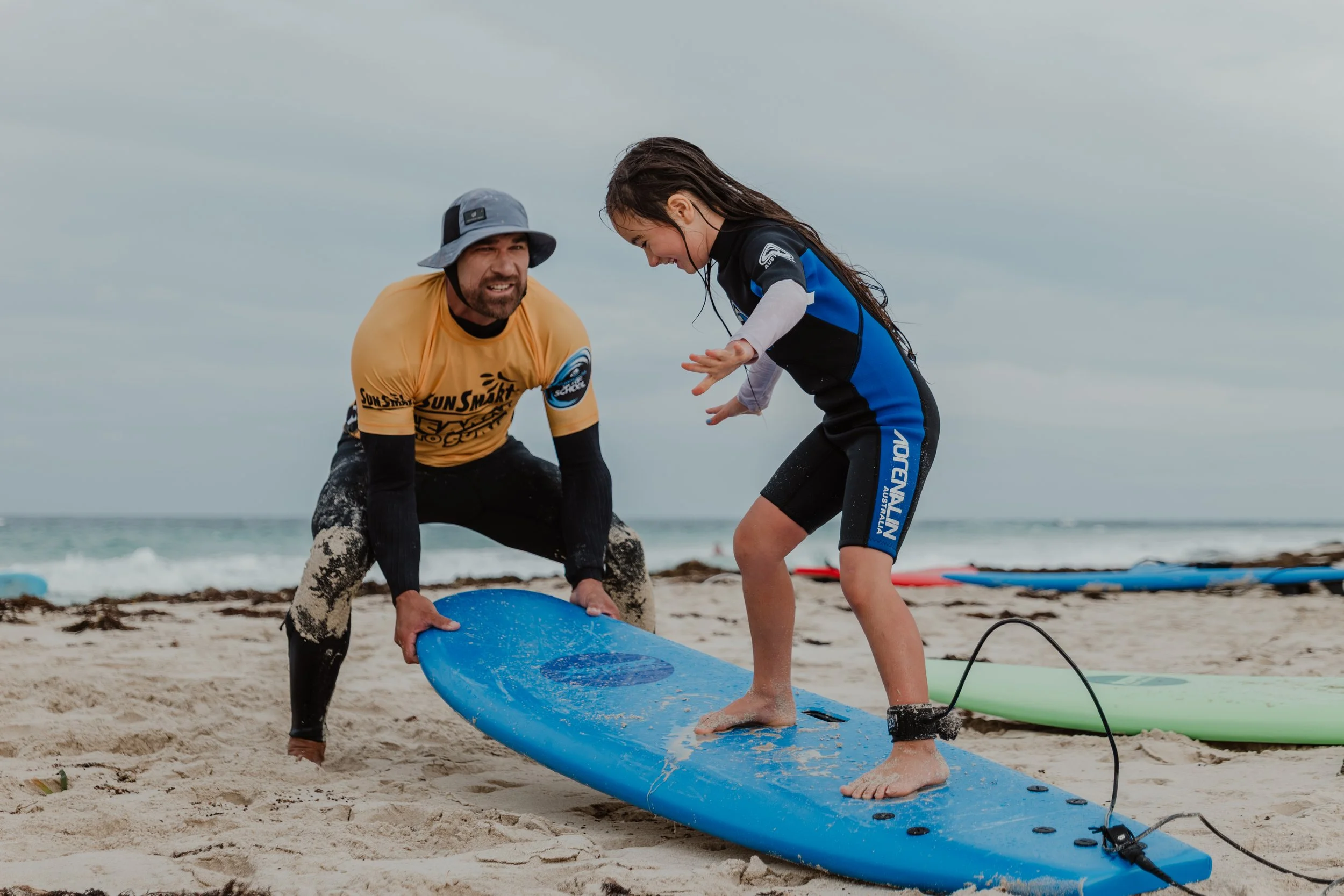 A young girl practicing surfing on her blue surfboard with the help of an instructor at the beach on a cloudy day, with other surfboards and the ocean in the background.