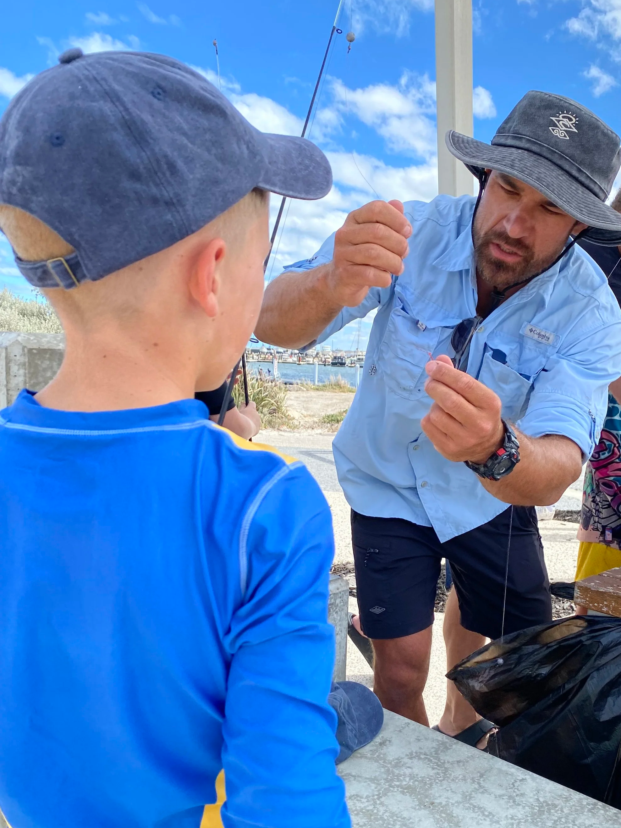 A man showing a fishing hook to a young boy outdoors near a body of water on a partly cloudy day.
