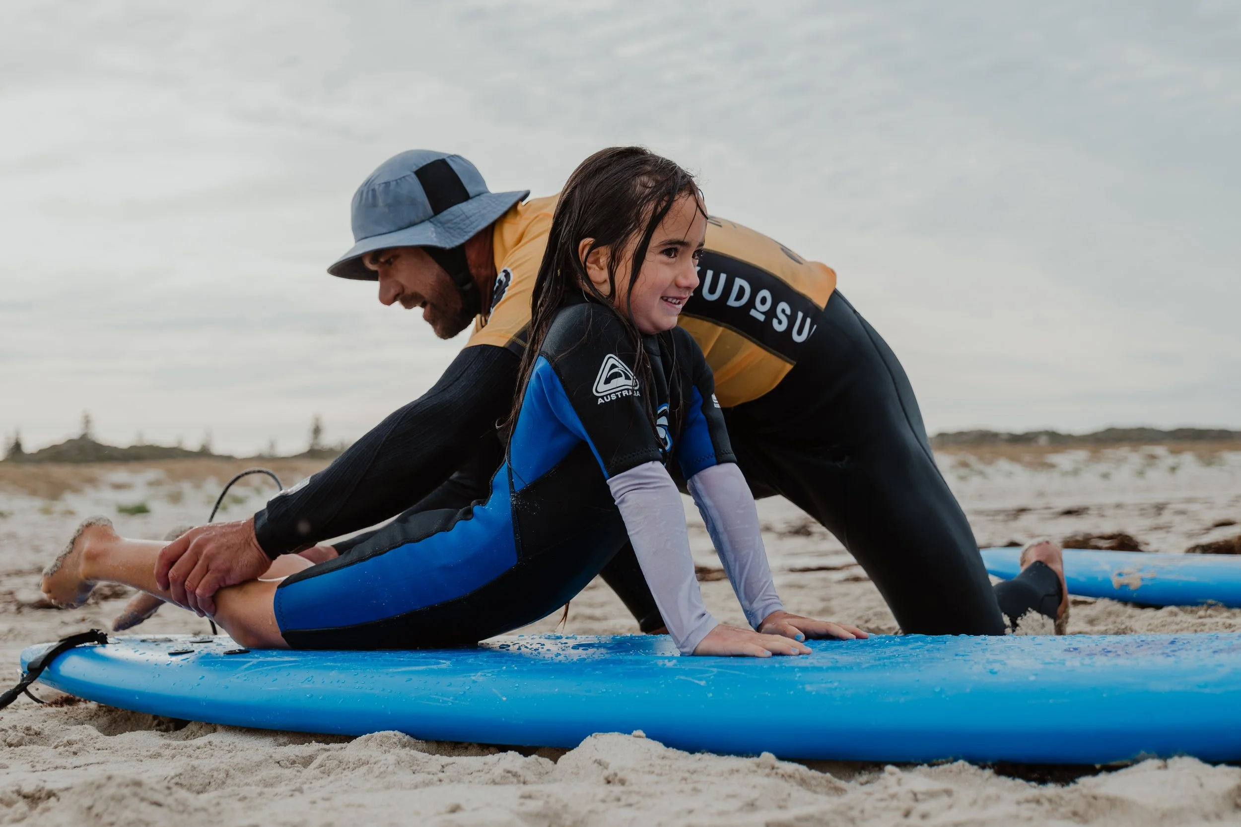 A man and a young girl practicing surfing on a sandy beach, both on a blue surfboard, with the girl in a wetsuit and the man assisting her.