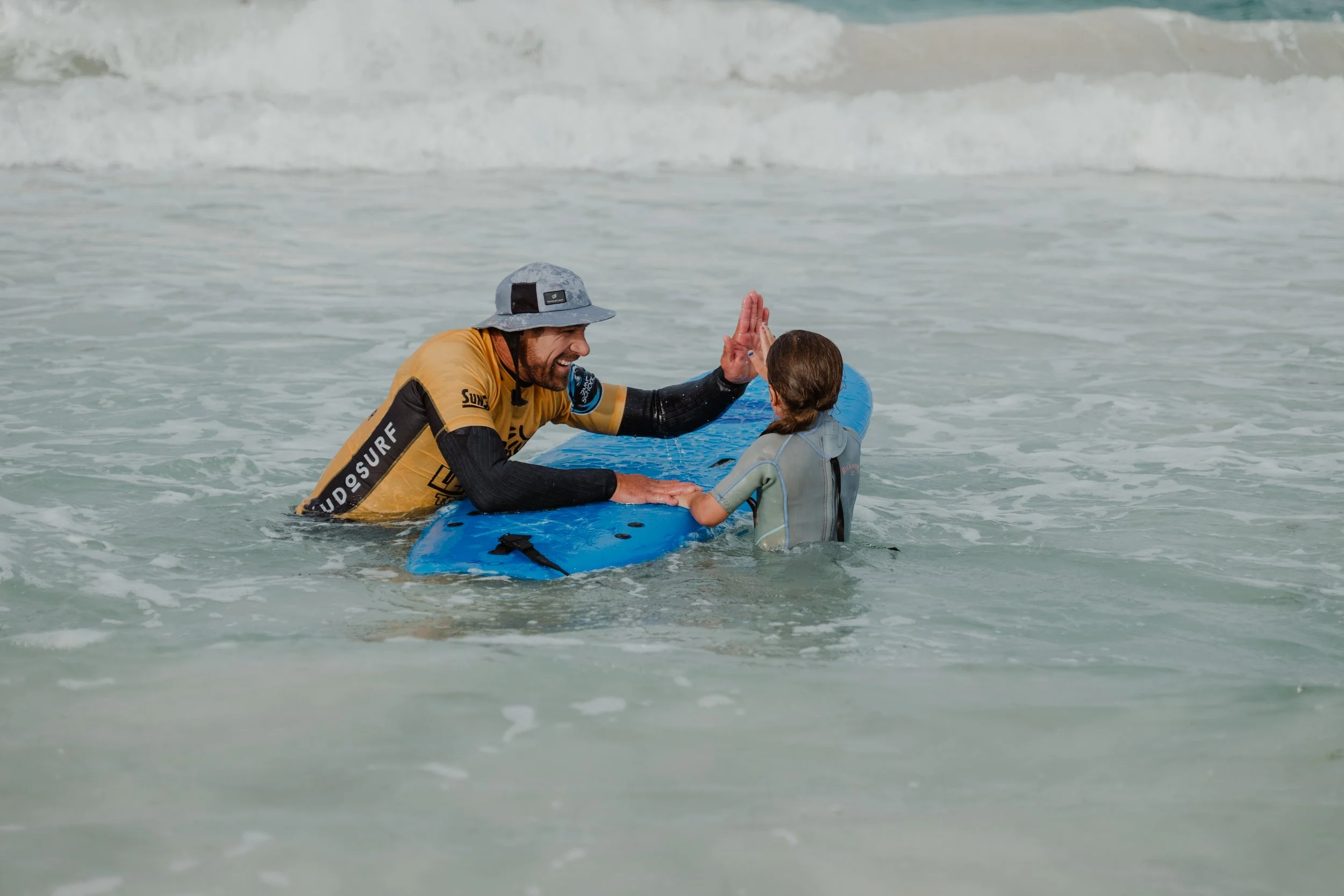A man in a yellow and black surfing shirt giving a high five to a young girl in a wetsuit while in the ocean, with a blue surfboard nearby.