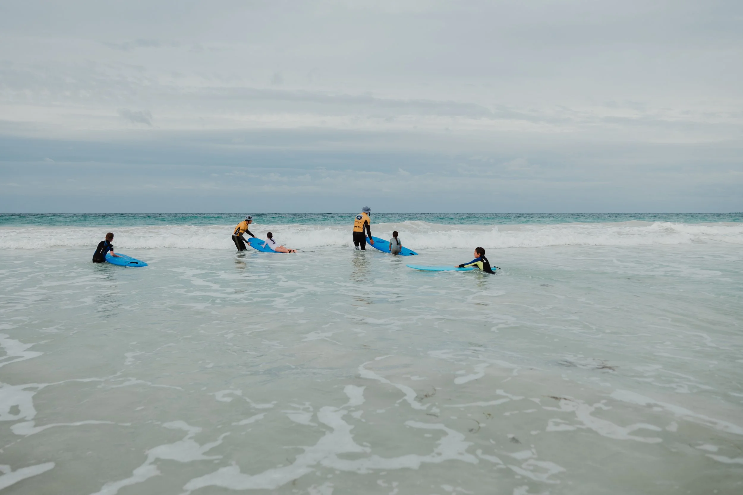 People, including children, in the ocean helping each other with surfboards during a surf lesson, with waves and cloudy sky in the background.