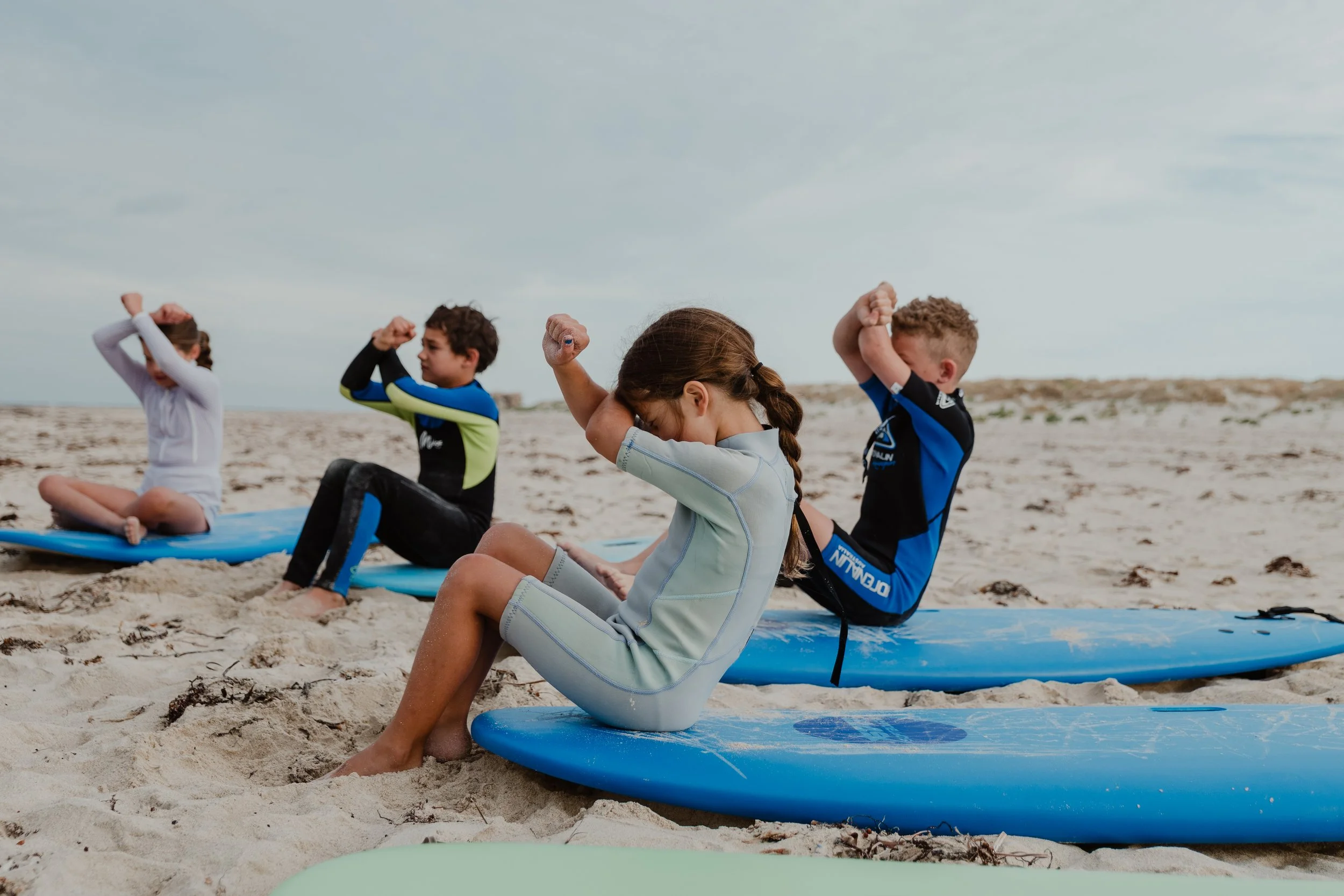 Four children sitting on surfboards on the sand at the beach, practicing yoga or meditation, with a cloudy sky in the background.