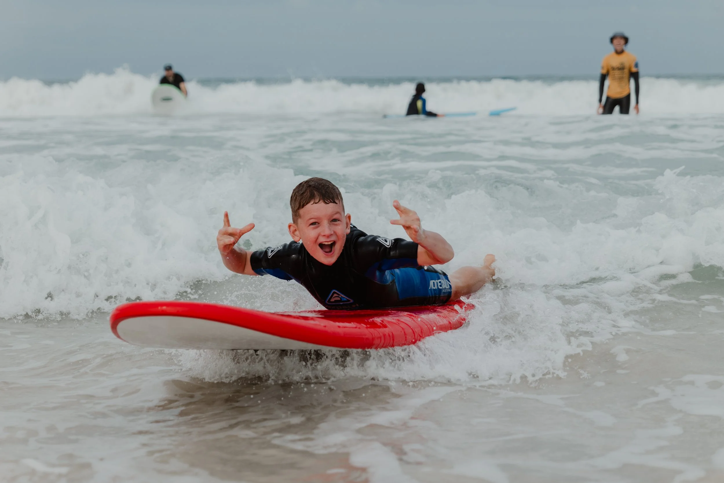 A young boy lying on a red surfboard, smiling and posing with his hands making peace signs while riding a wave at the beach with other surfers and people in the background.