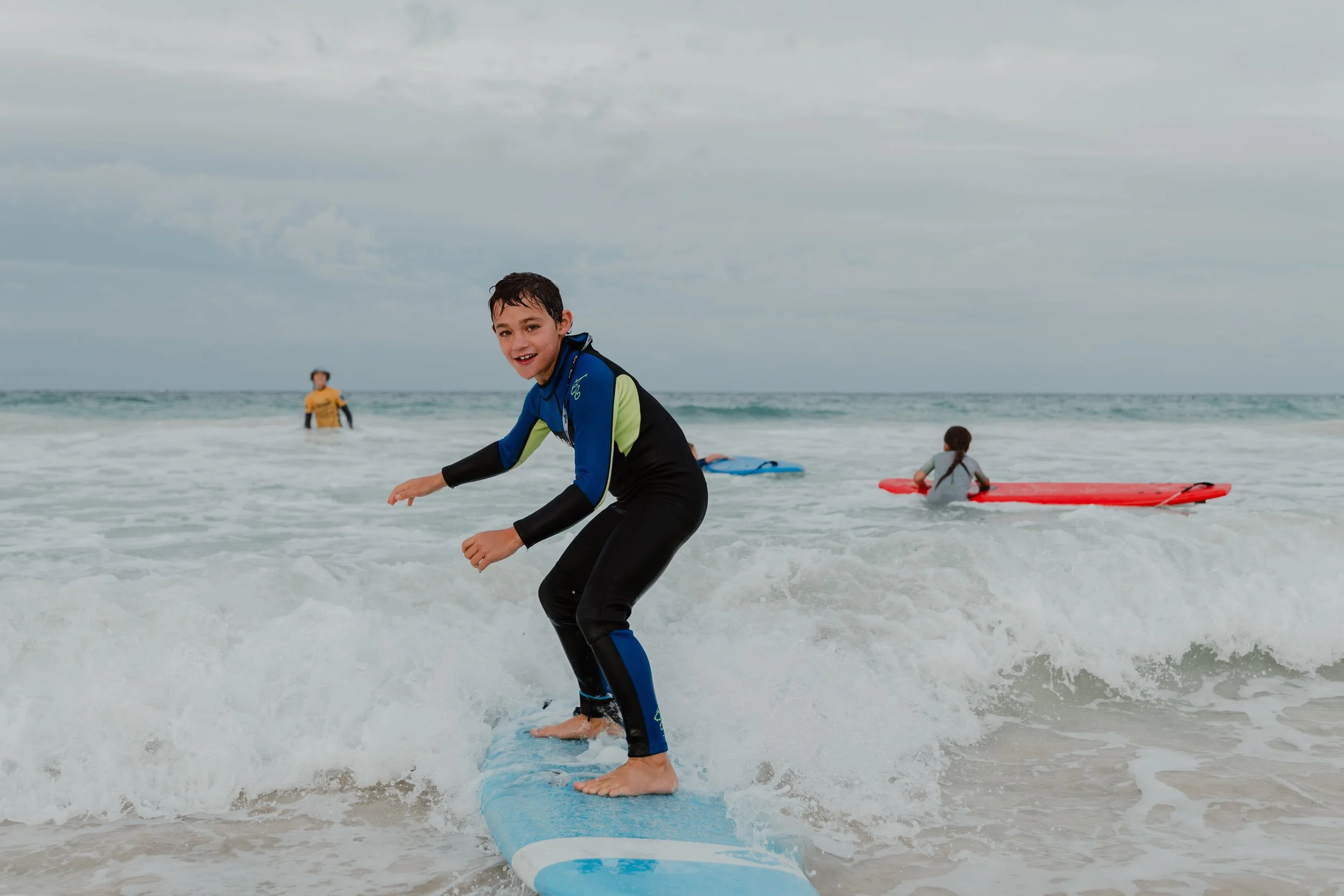 A boy surfing on a blue surfboard in the ocean with two other children in the background, one on a red surfboard and another in the water with a yellow shirt, under a cloudy sky.