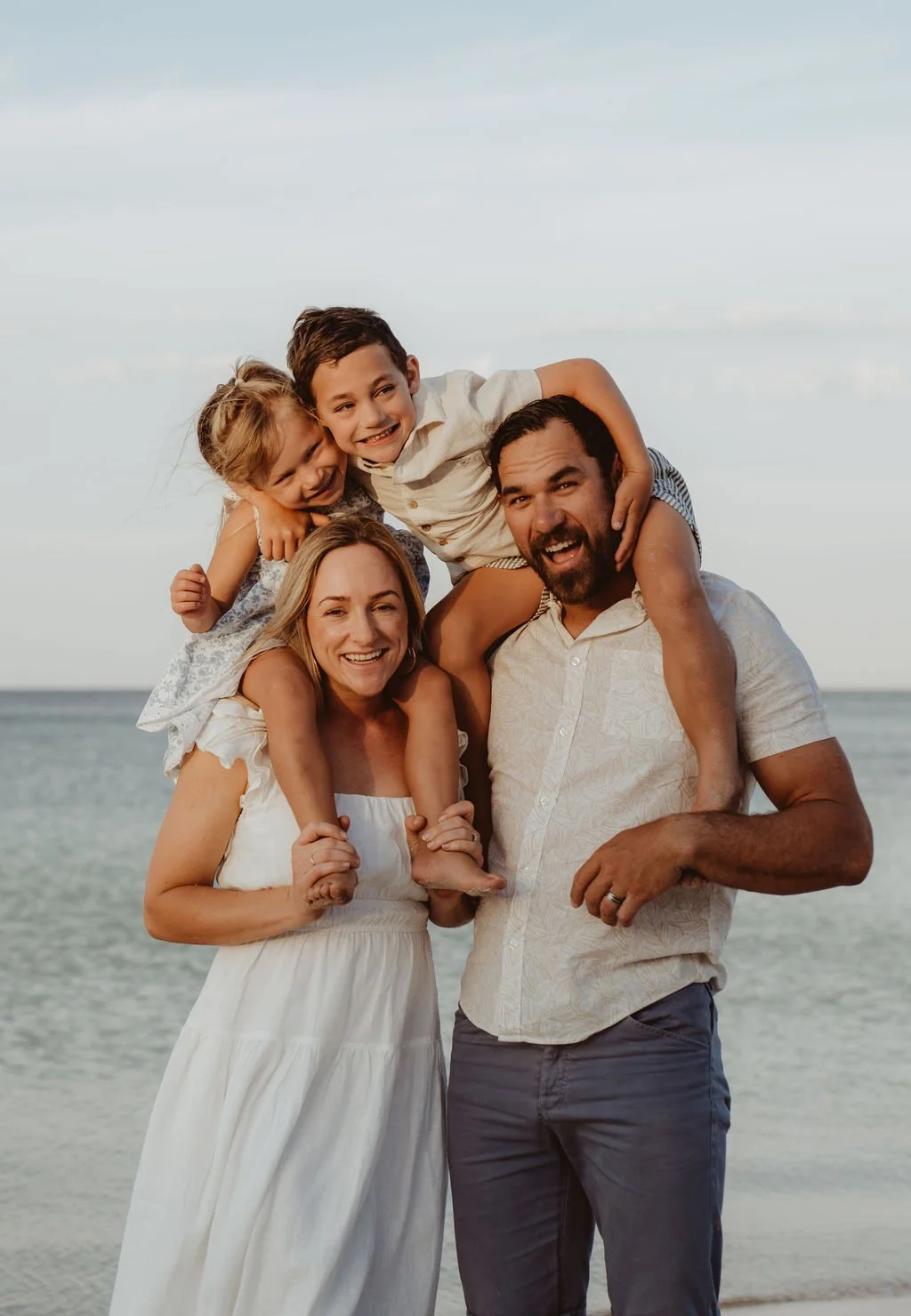 A family of four at the beach, with the father carrying a boy on his shoulders and the mother holding a girl, all smiling.