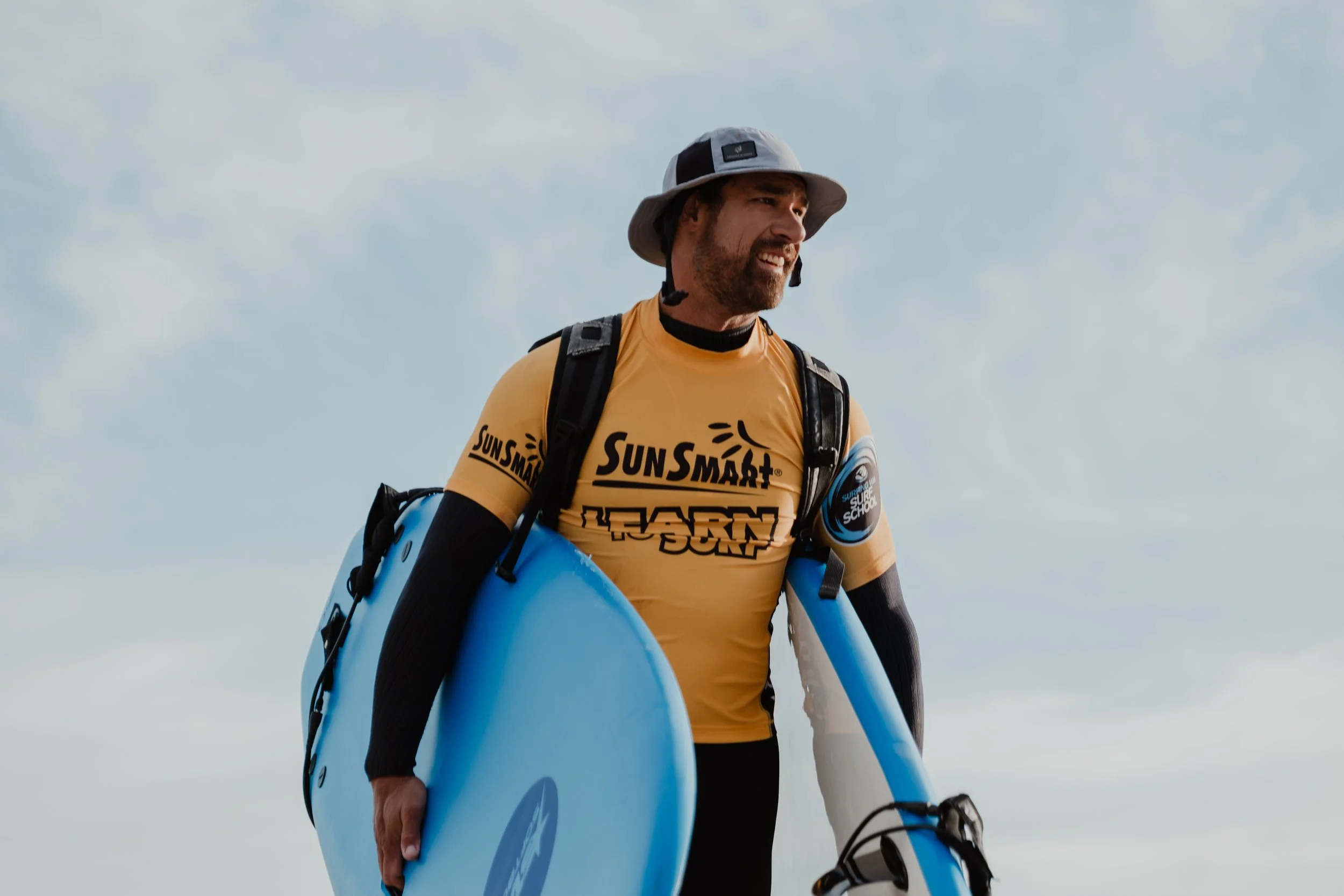 Man in yellow SunSmart shirt holding a blue surfboard on a beach with cloudy sky.