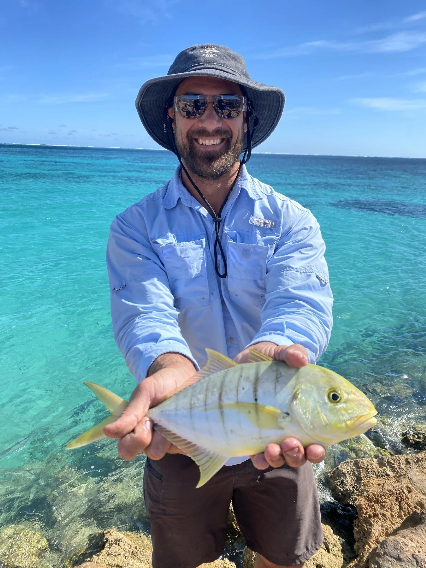 A man wearing a wide-brimmed hat, sunglasses, and a long-sleeved shirt holds a fish in front of a clear blue ocean background.