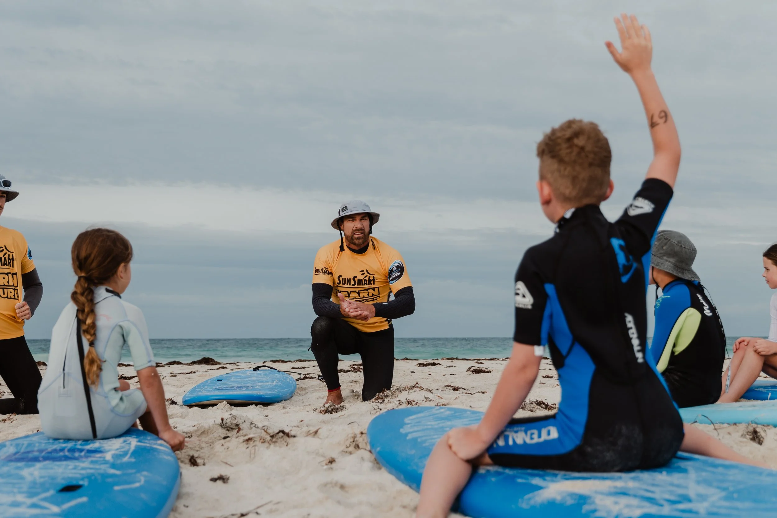 A surf instructor kneeling on the sand, giving a lesson to children seated on surfboards on the beach, with the ocean and cloudy sky in the background.