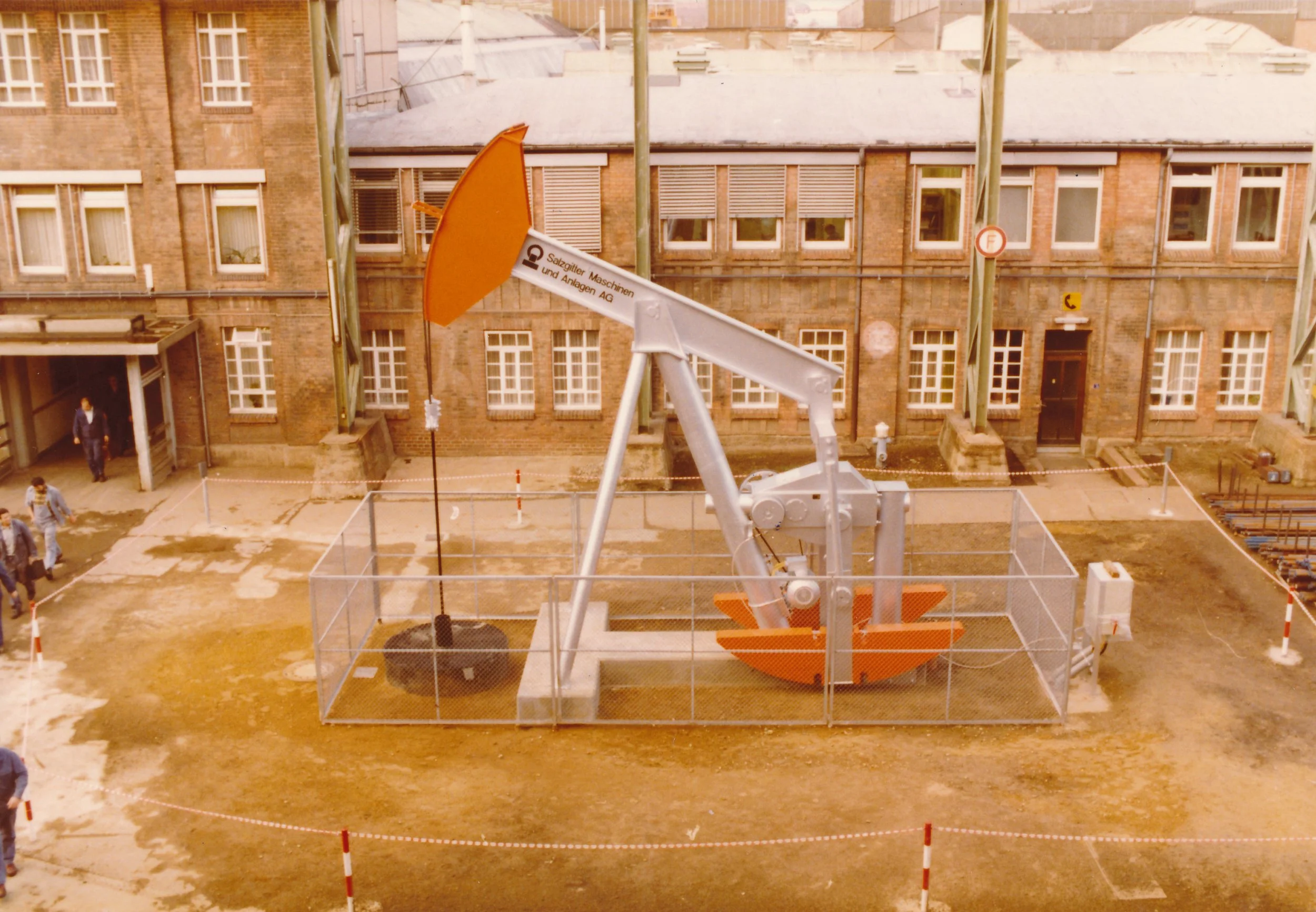 Bauarbeiter und Baustelle mit einer großen Baukräne in der Mitte, umgeben von einem Sicherheitszaun, im Hintergrund ein altes mehrstöckiges Backsteingebäude.