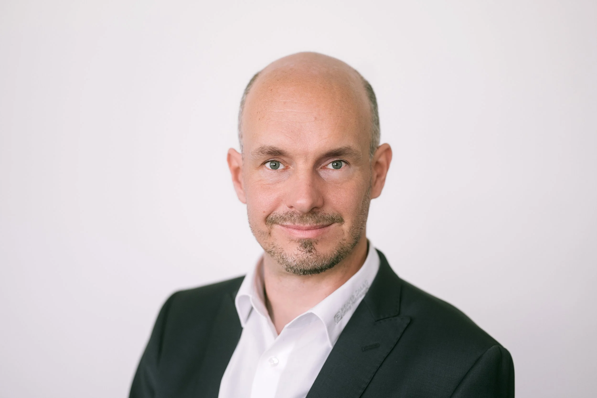 Porträt eines Mannes mit Glatze, blauen Augen, Bart und gepflegtem Gesichtsausdruck, trägt einen schwarzen Anzug und weißes Hemd vor weißem Hintergrund.