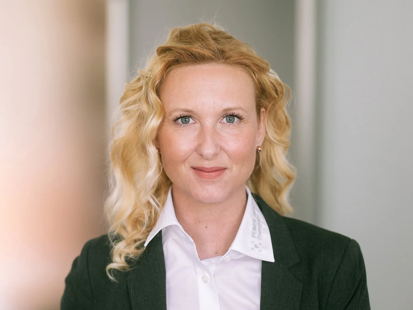 Porträt einer Frau mit blonden, lockigen Haaren, weißem Hemd und dunklem Blazer vor einer neutralen Wand.