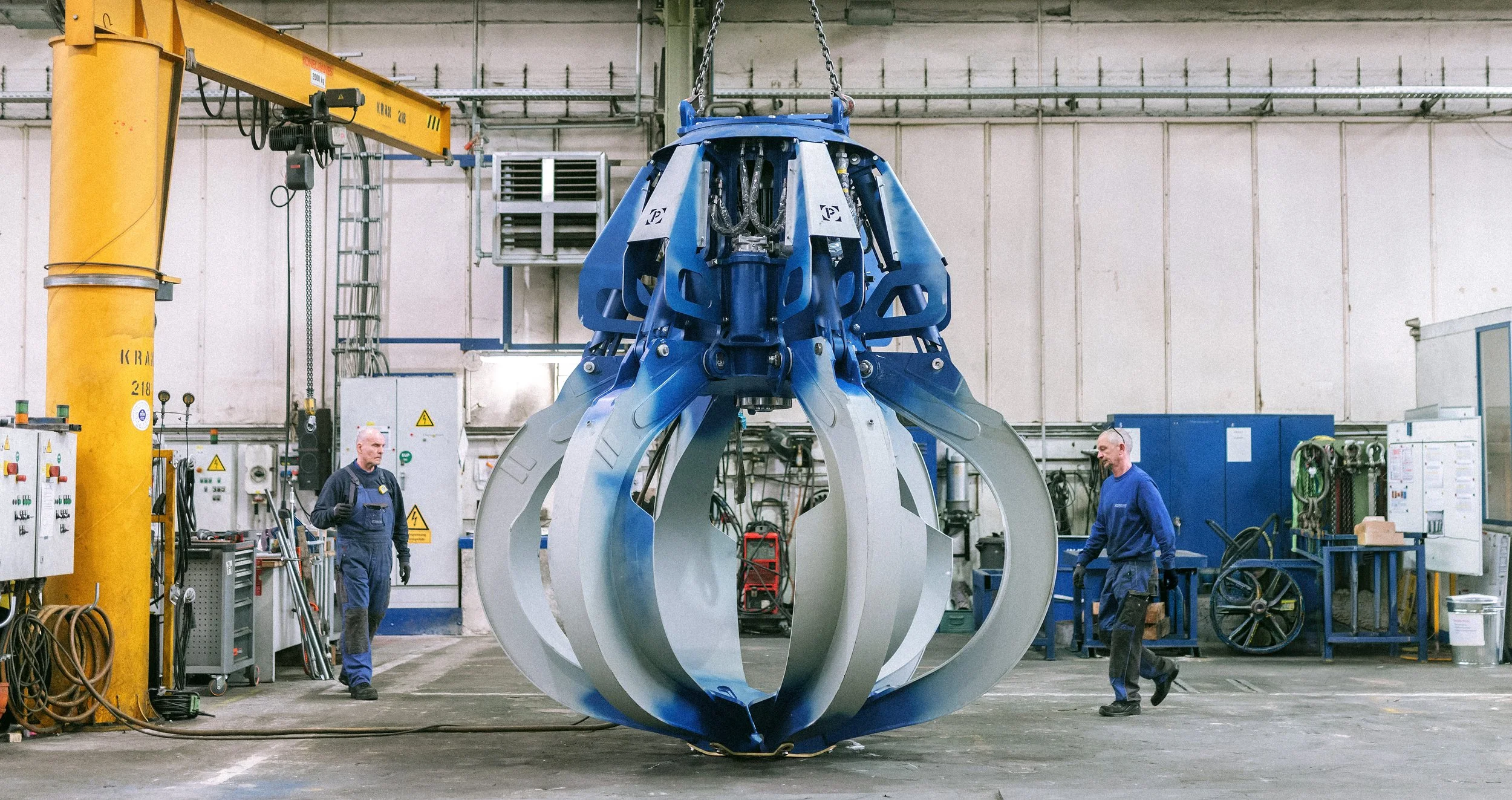 Zwei Facharbeiter arbeiten in einer Fabrik an einem großen mechanischen Teil, das in der Mitte einer Werkstatt hängt, neben einem gelben Kran.