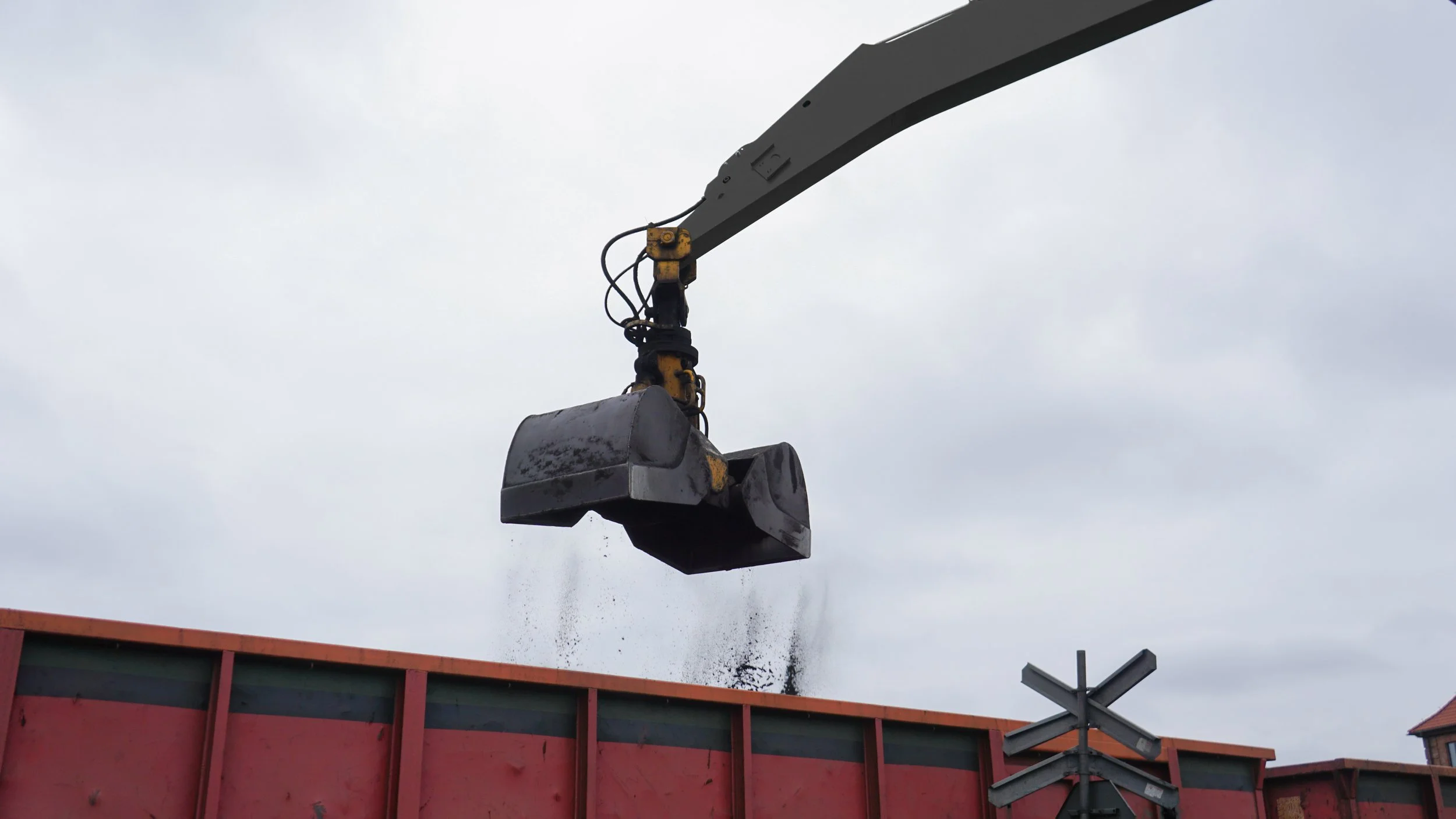 Ein Kran schwingt eine große Metallkiste in die Luft, während Räumpulver oder Staub vom Boden aufwirbelt, bei bewölktem Himmel.
