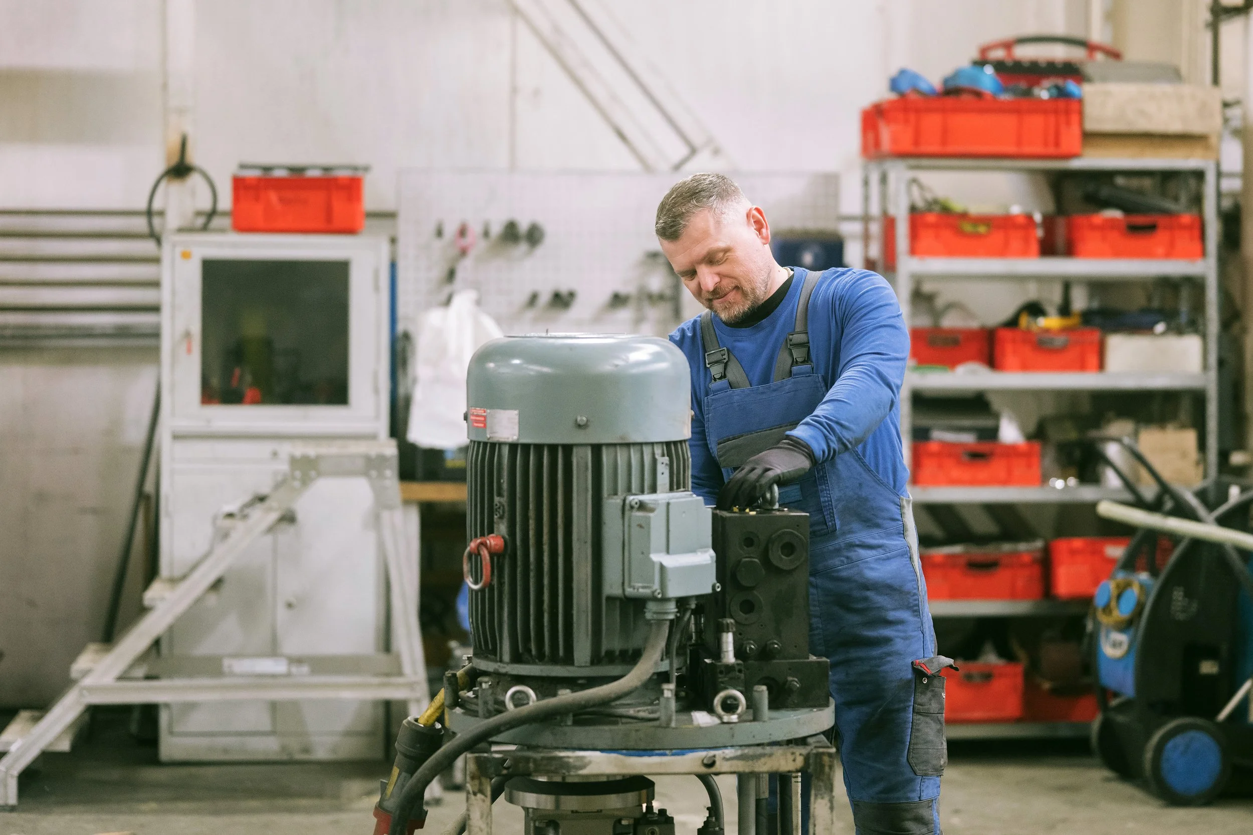 Ein Mann arbeitet in einer Werkstatt an einer Maschine, umgeben von Werkzeugen und Werkstoffen, mit roten Kisten im Hintergrund.