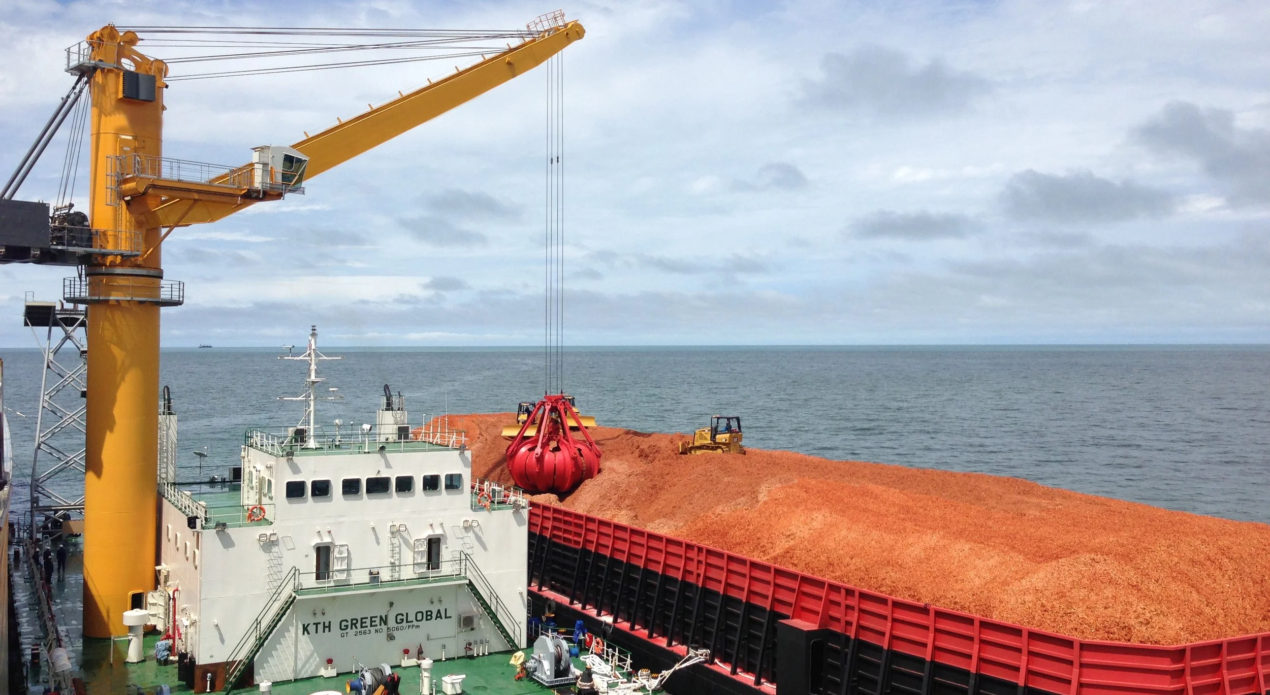Ein offenes Schiffsfrachtterminal mit großen Ladungscontainern, einem Kran, der mit einem roten Fangkorb eine Ladung auf ein Schiff hebt, im Hintergrund das Meer und ein Himmel mit Wolken.