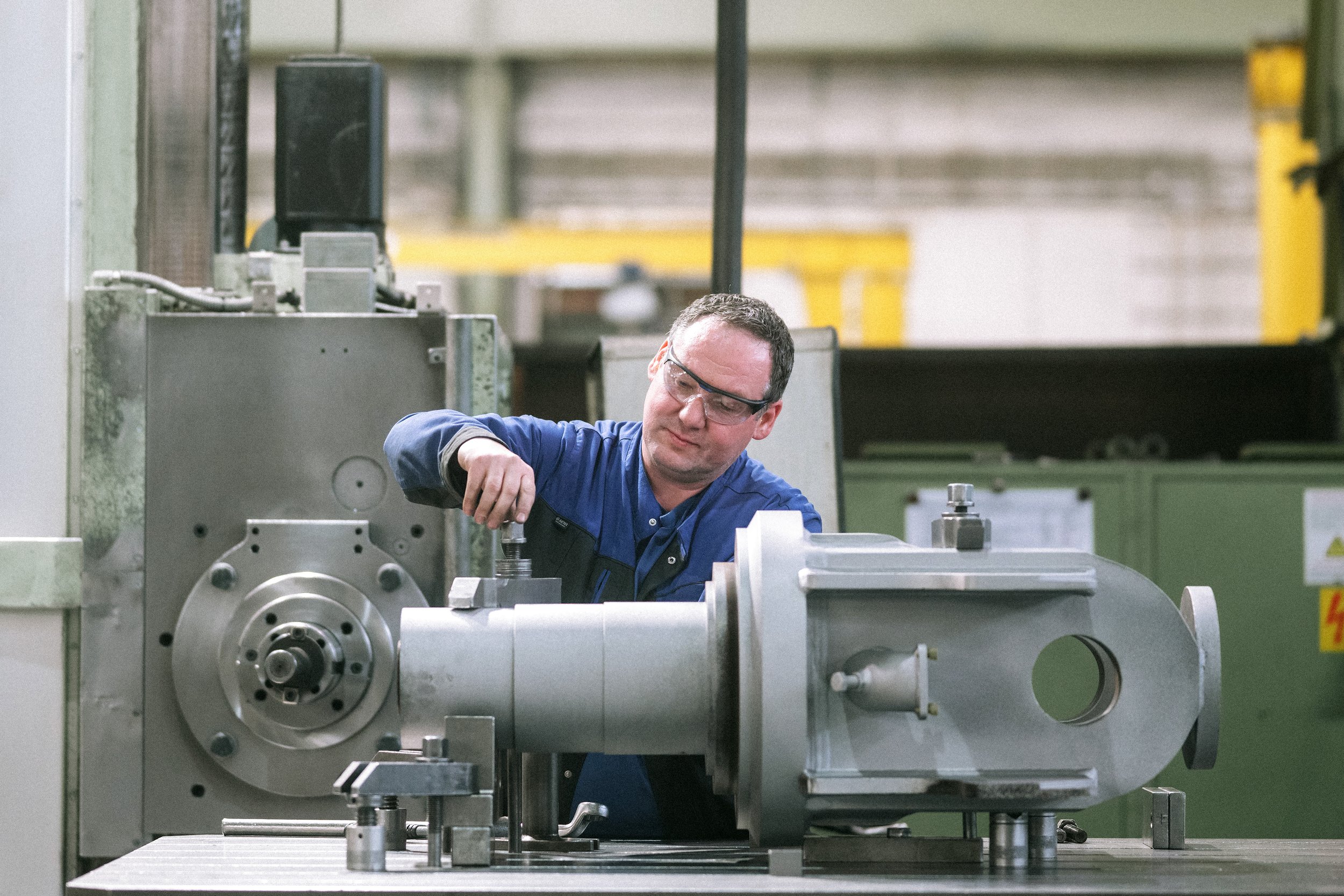 Ein Mann arbeitet in einer Fabrik an einer Maschine, trägt Schutzbrille und blaue Arbeitskleidung.