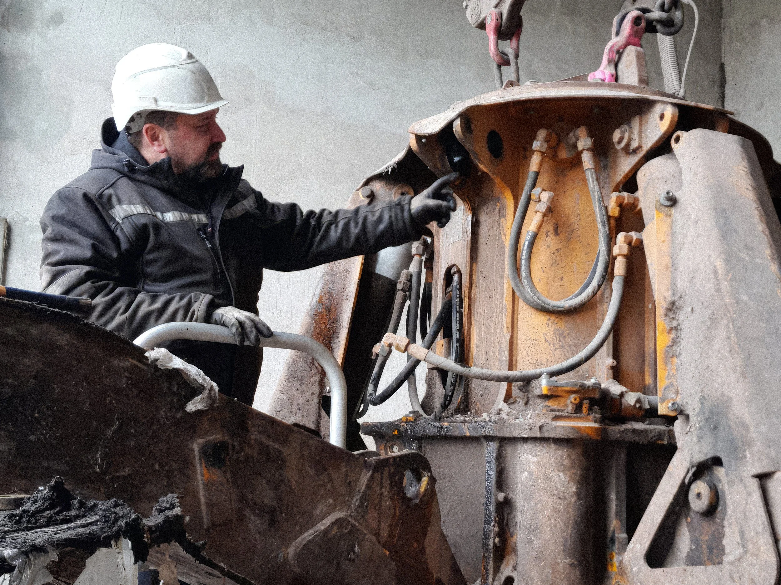 Ein Bauarbeiter trägt einen weißen Schutzhelm und Arbeitskleidung, während er an einem großen, rustikalen Maschinen- oder Maschinenteil arbeitet, das mit verschiedenen Rohren und Schrauben verbunden ist.