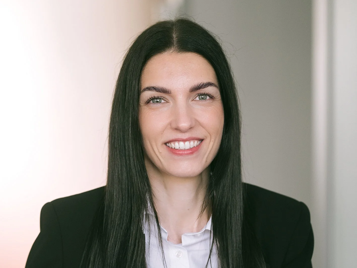 Ein Porträt einer lachenden Frau mit langen, glatten schwarzen Haaren, weißem Hemd und dunklem Blazer vor einem neutralen Hintergrund.